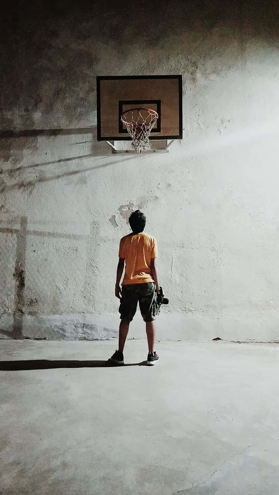Basketball Aesthetic 桌布 壁紙 — 一個身穿橙色襯衫的孤獨籃球運動員站在光線昏暗的混凝土球場上一個破舊的籃框下。