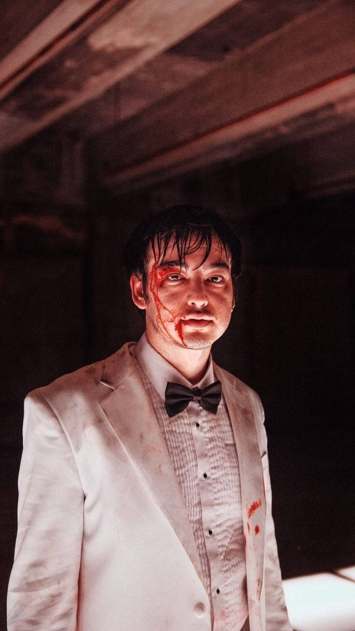 joji 桌布 壁紙 — 一個身穿沾滿血跡的白色西裝的男子，在一個光線昏暗、腐朽的房間裡，木樑下凝視著。