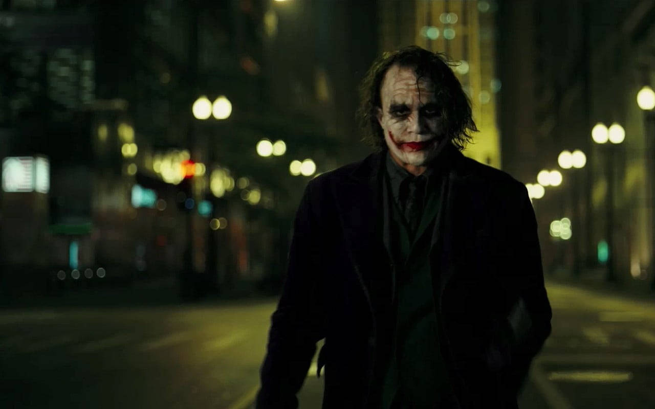 Heath Ledger Joker 桌布 壁紙 — 一個身穿深色外套、帶有威脅性的小丑在夜間走在被雨水浸濕的城市街道上，背景有發光的街燈。