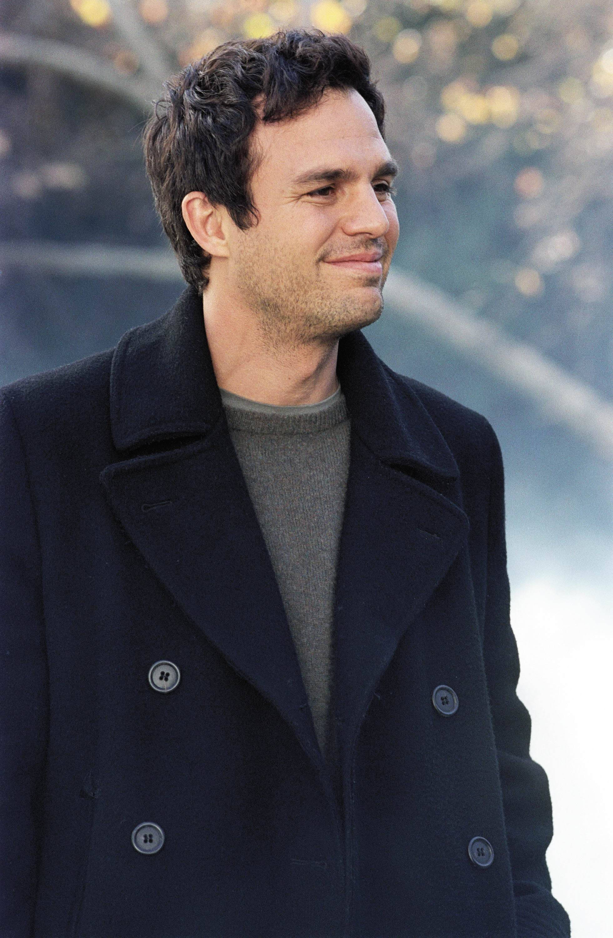 Mark Ruffalo 桌布 壁紙 — 一個身穿深色外套的男人站在戶外，背景有模糊的秋葉和水面，溫柔地微笑。