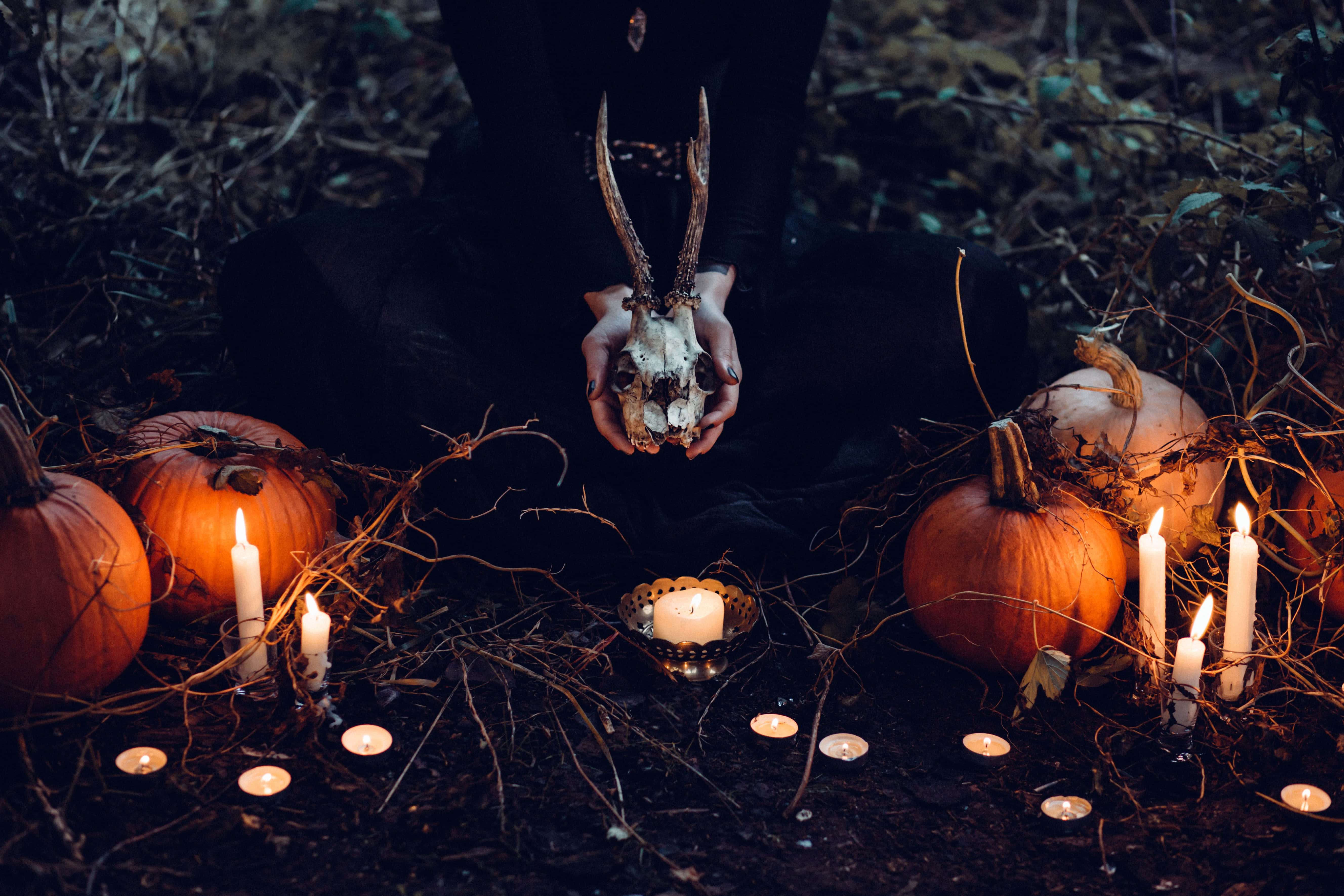 Spooky Halloween 桌布 壁紙 — 一個身穿深色衣服的人，在昏暗的森林中，手持帶鹿角的鹿頭骨，周圍環繞著發光的蠟燭和南瓜。