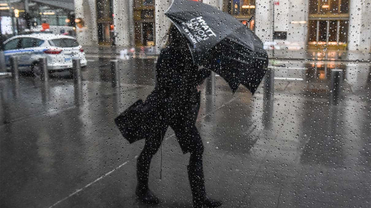 下雨 桌布 壁紙 — 一個身穿深色衣服的人撐著一把有白色文字的黑色雨傘，在雨中的城市街道上快步行走，前景玻璃上有水滴劃過的痕跡。