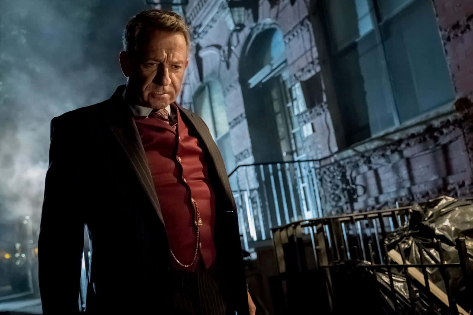 Alfred Pennyworth 桌布 壁紙 — 一個身穿深色西裝和紅色背心的男人，夜晚站在煙霧瀰漫的城市小巷中，表情嚴肅。