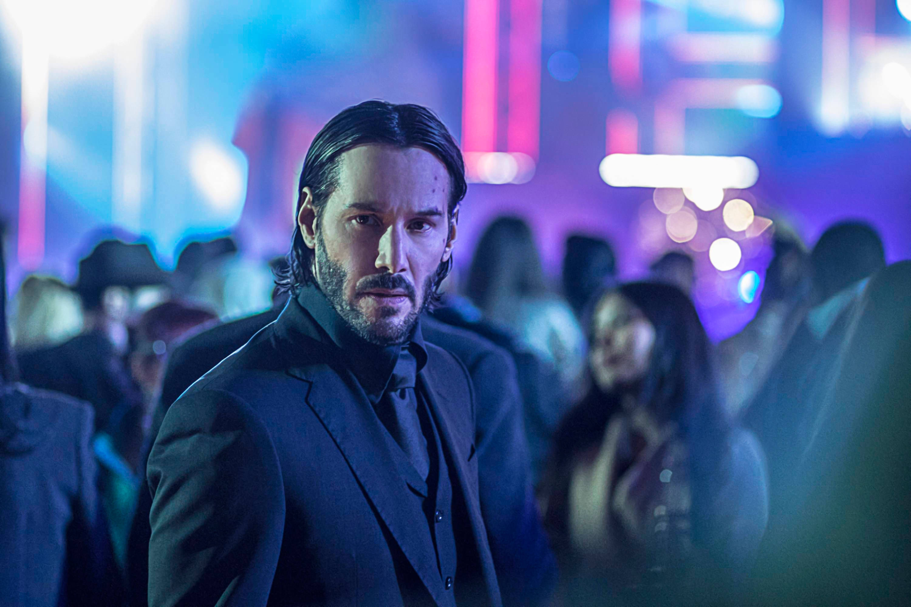 John Wick 桌布 壁紙 — 一個身穿深色西裝的男人在霓虹燈光下的人群中凝視，人群中有模糊的身影和鮮豔的藍色和粉紅色燈光。