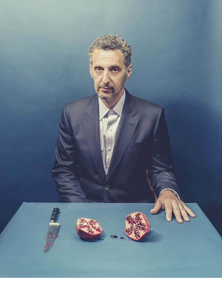 John Turturro 桌布 壁紙 — 一個身穿深色西裝的男人坐在桌前，桌上放著切開的石榴和一把刀，背景為藍色，他正凝視著鏡頭。