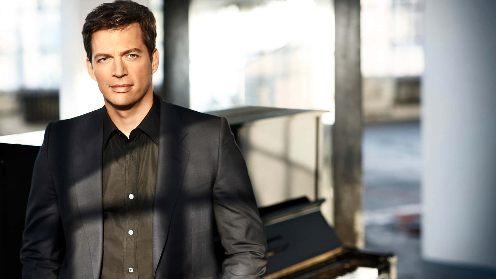 Harry Connick Jr 桌布 壁紙 — 一個身穿深色西裝的男人自信地站在現代、陽光充足的房間裡，旁邊有一架三角鋼琴，房間有大窗戶。