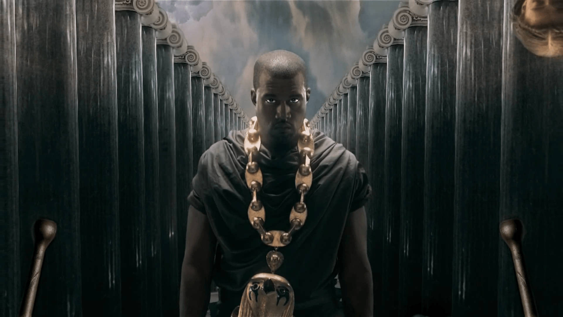 Kanye 桌布 壁紙 — 一個身穿深色長袍、戴著沉重金鏈的男人，在暴風雨的天空下，莊嚴地站在高聳華麗的柱子之間。