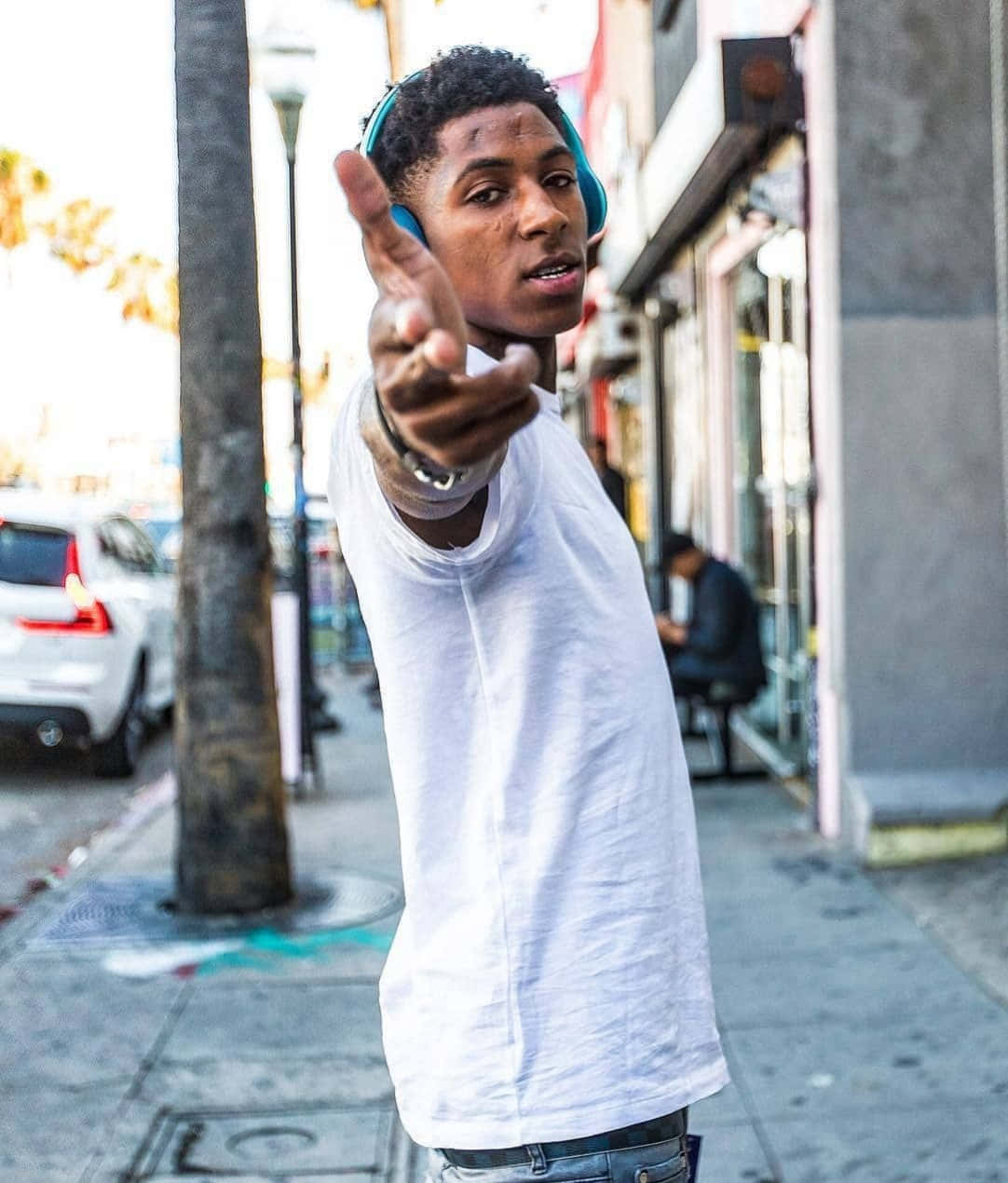 NBA YoungBoy 桌布 壁紙 — 一個身穿白色T恤和藍色耳機的年輕男子在陽光明媚的城市人行道上向鏡頭伸出手。