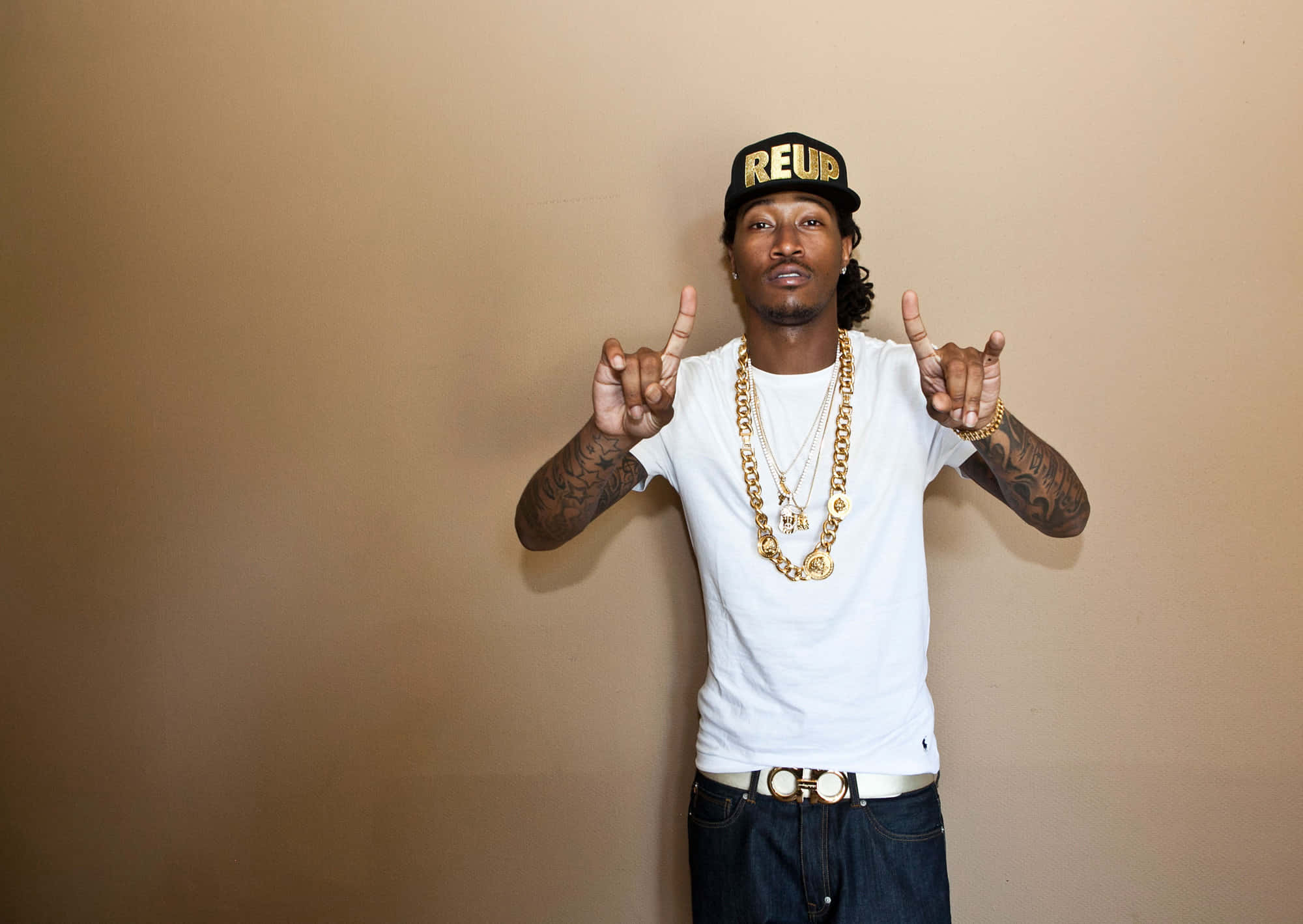 Rappers 桌布 壁紙 — 一個身穿白色T恤和金色鏈條的男人，在純色米色牆前自信地擺姿勢，雙手做出搖滾手勢。