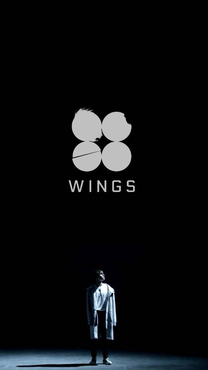 Bts Black 桌布 壁紙 — 一個身穿白色外套的孤獨身影，在黑暗中站在一個帶有四個圓形元素的發光「WINGS」標誌下方。