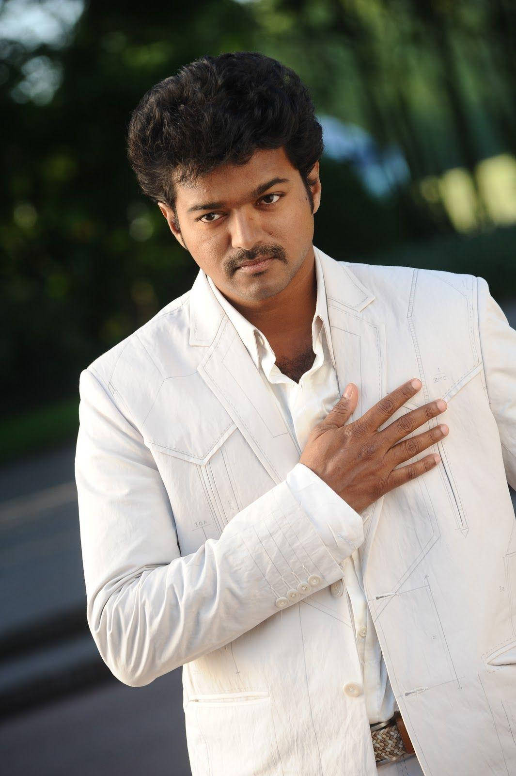 vijay HD 桌布 壁紙 — 一個身穿白色西裝的男人自信地站著，手放在胸前，背景是模糊的綠色戶外景色。