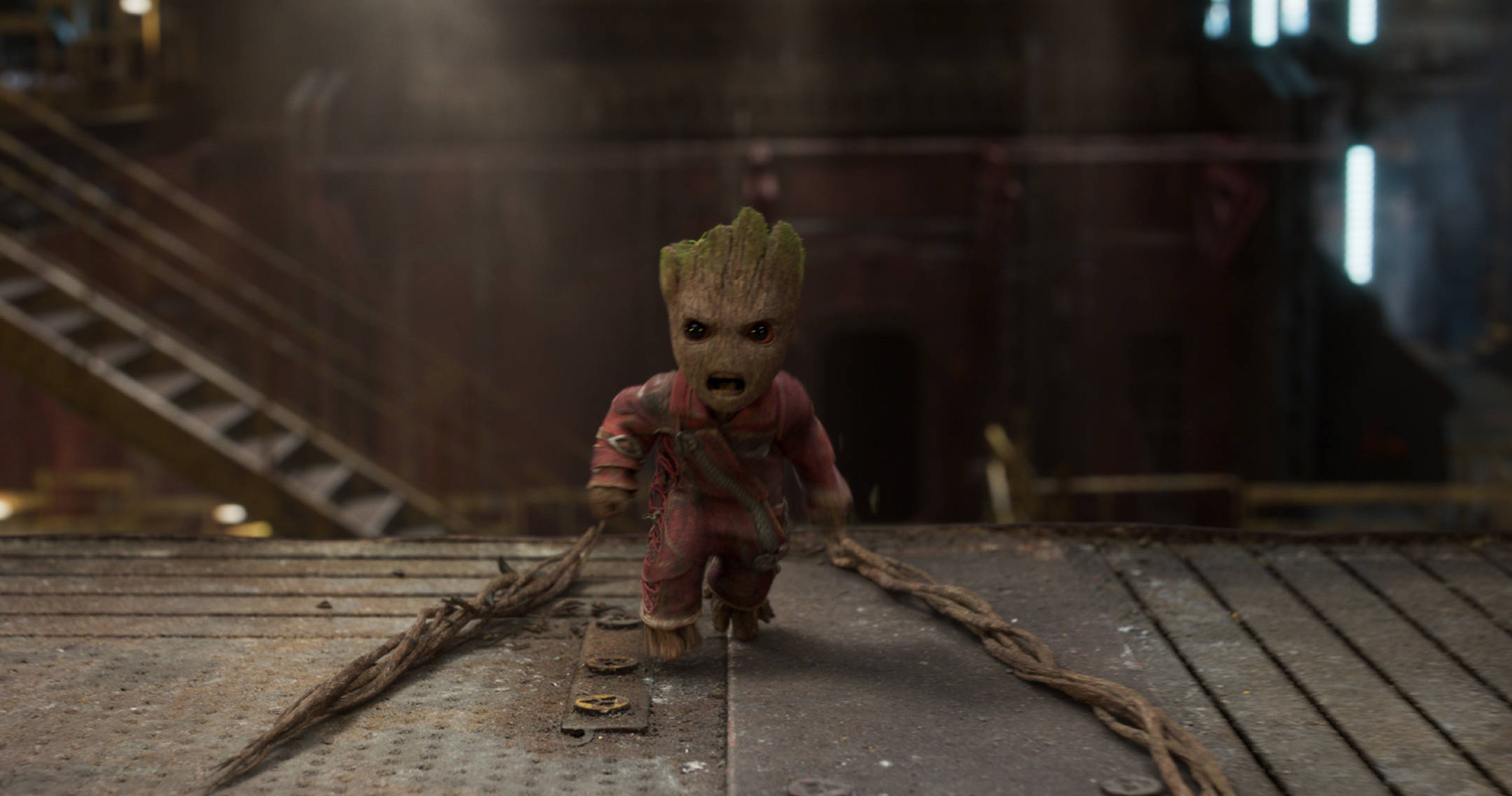 Baby Groot 桌布 壁紙 — 一個身穿紅色服裝的小格魯特站在金屬格柵上，藤蔓在昏暗的工業環境中拖在身後。