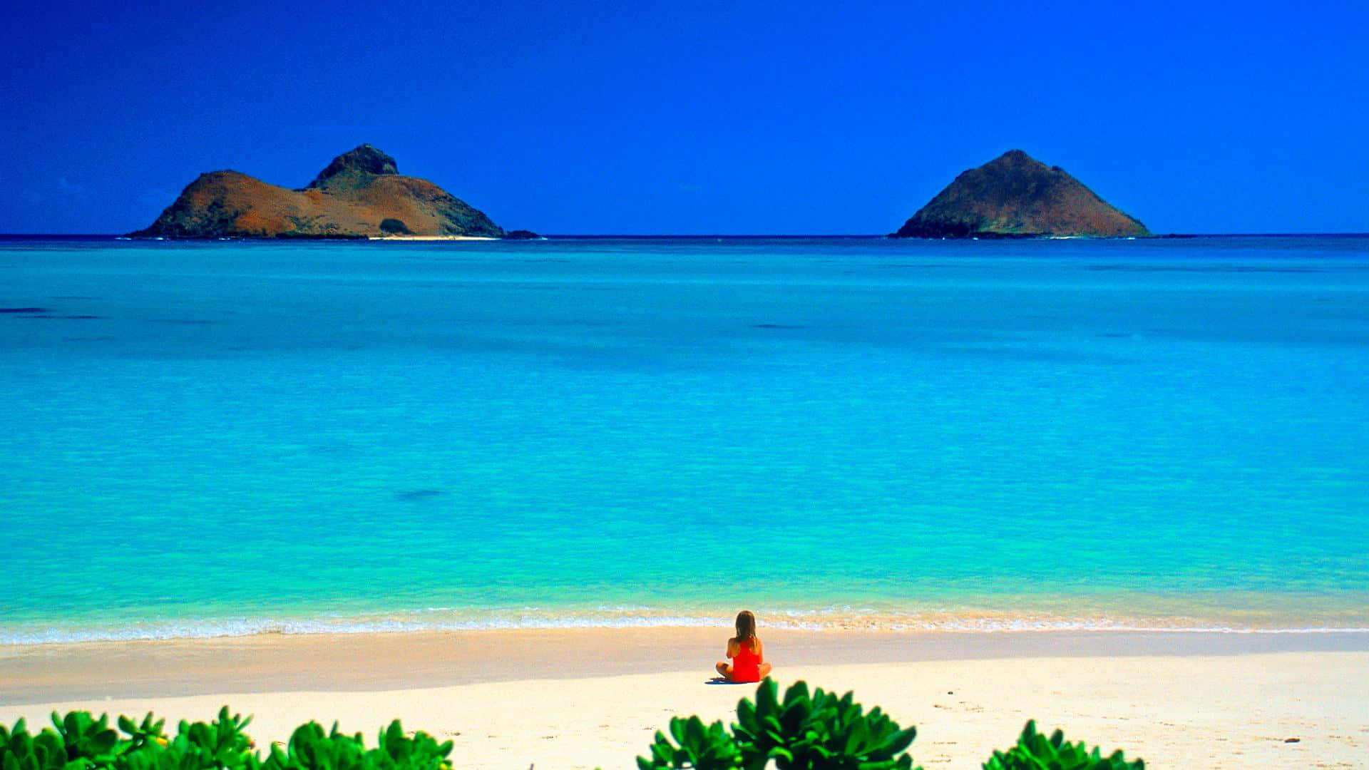 Hawaiian Beach 桌布 壁紙 — 一個身穿紅色衣服的人獨自坐在純淨的白色沙灘上，面向碧綠的海水和遠處的火山島嶼，天空湛藍。