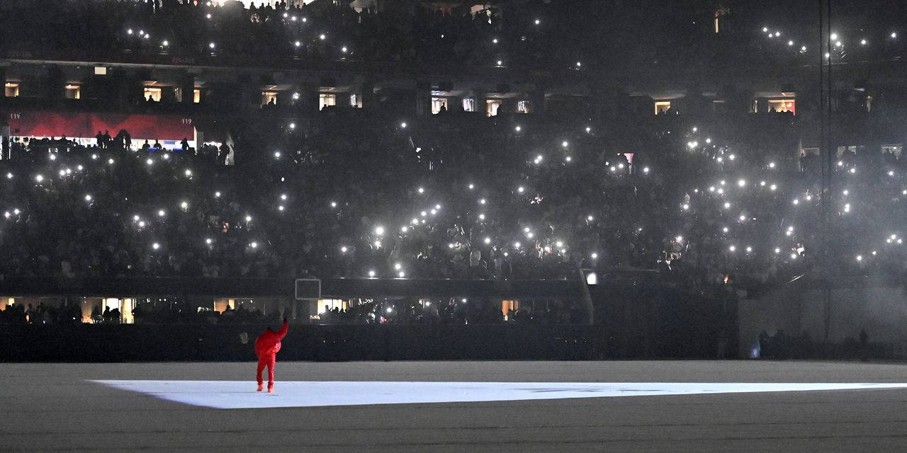 Kanye West Donda 桌布 壁紙 — 一個身穿紅色衣服的孤獨人物站在舞台中央，在明亮的燈光下，體育場看台上的人群舉起發光的手機螢幕。