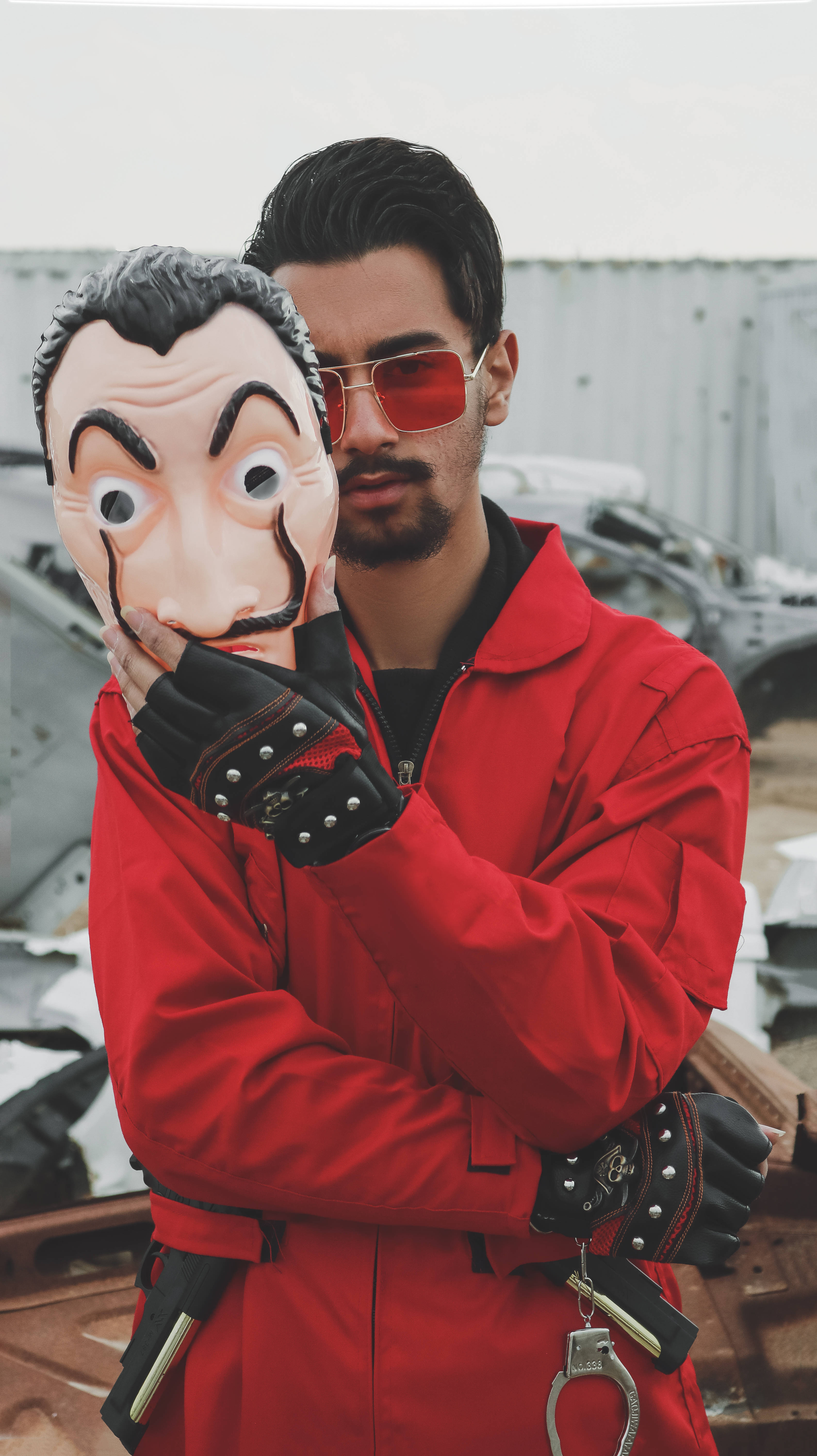 Money Heist Mask 桌布 壁紙 — 一個身穿紅色連身衣和紅色太陽眼鏡的男人，站在廢車場，手持薩爾瓦多·達利面具。