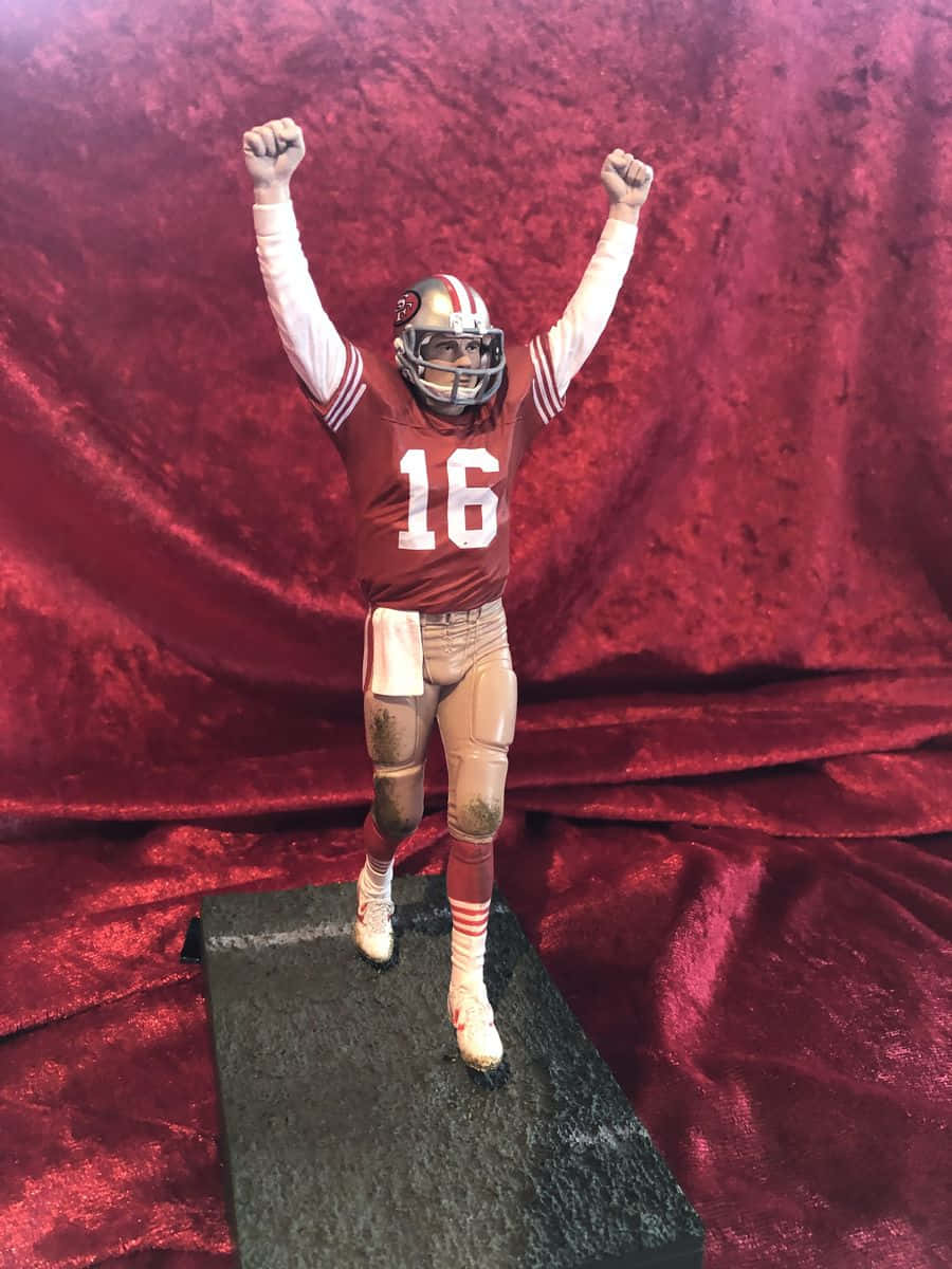 Joe Montana 桌布 壁紙 — 一個身穿紅金色球衣、號碼16的足球員人偶，在黑色底座上，以豐富的紅色天鵝絨背景襯托下，勝利地站立著。
