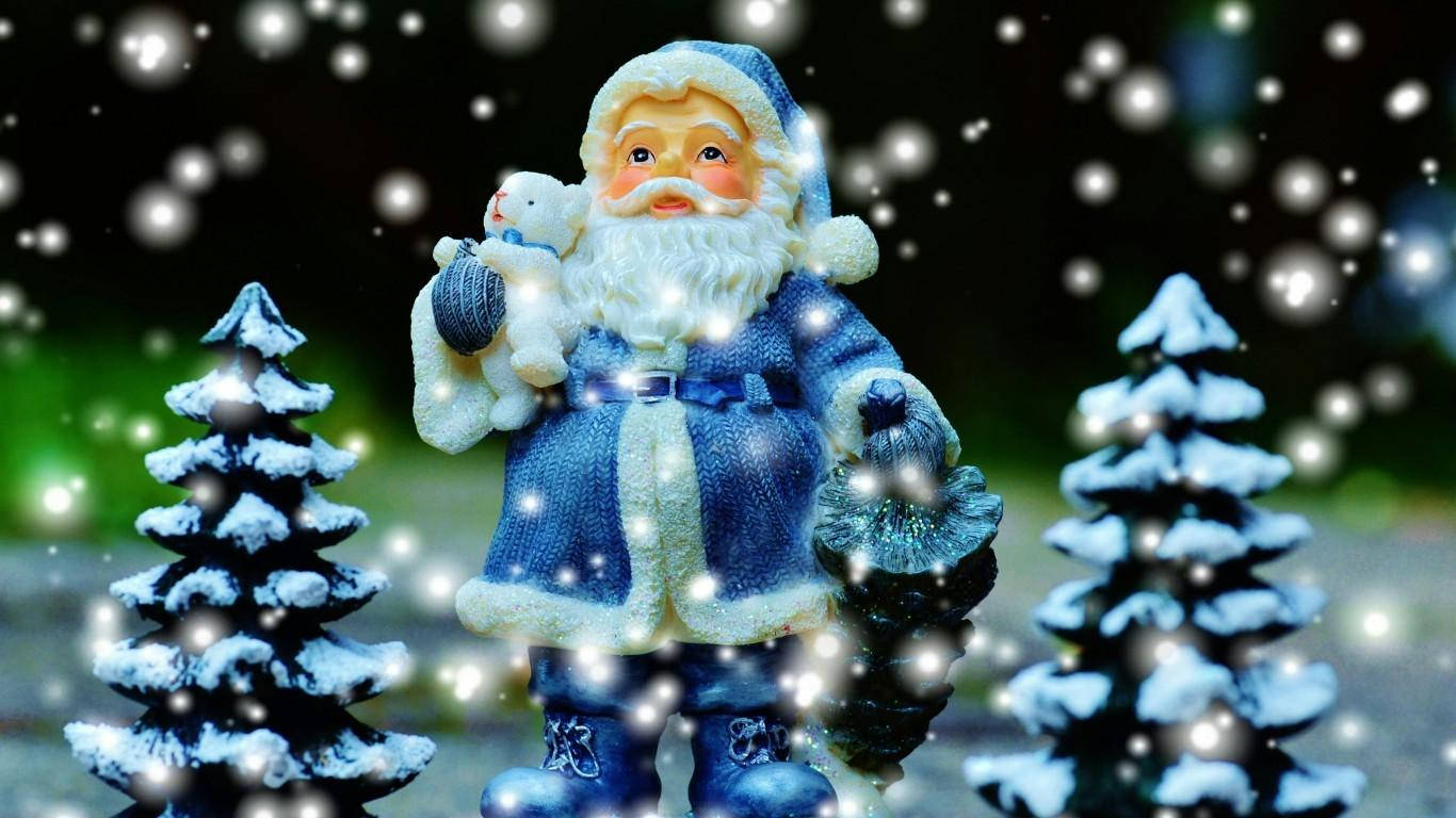 christmas laptop 桌布 壁紙 — 一個身穿藍色衣服的聖誕老人公仔在飄落的雪花和鋪滿雪的松樹之間抱著一隻泰迪熊。