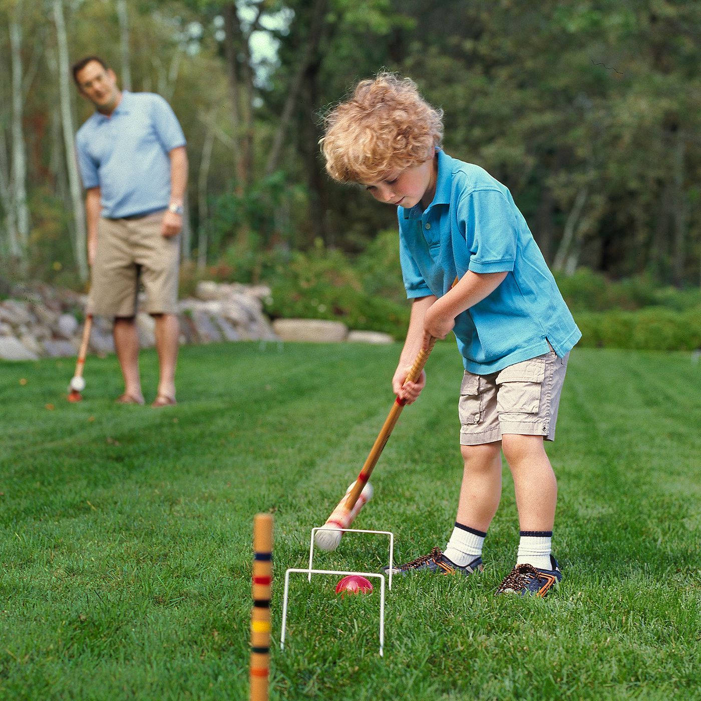 ball 桌布 壁紙 — 一個身穿藍色襯衫的小男孩專注地將槌球擊過球門，背景是一片翠綠的草坪，一名男子在旁觀看。