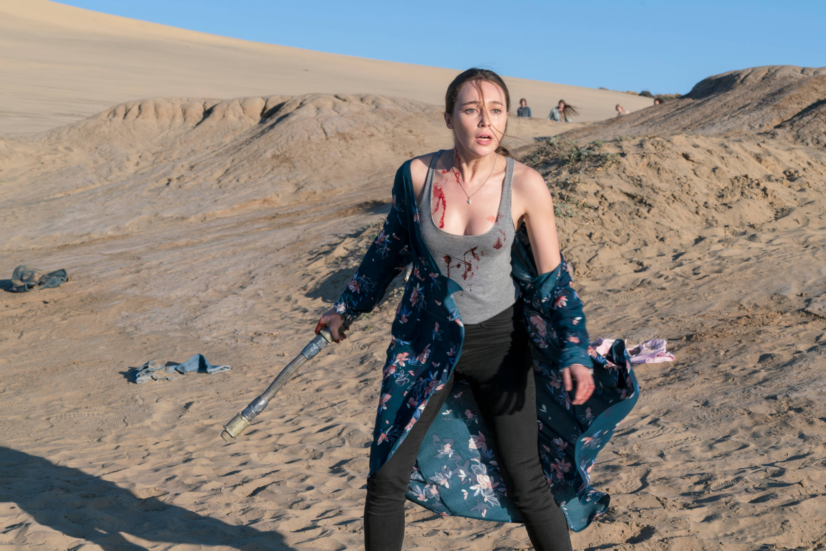 Desert 桌布 壁紙 — 一個身穿血衣的女人手持球棒，在陽光明媚的沙漠中，沙丘和遠處的人影中挑釁地站立。