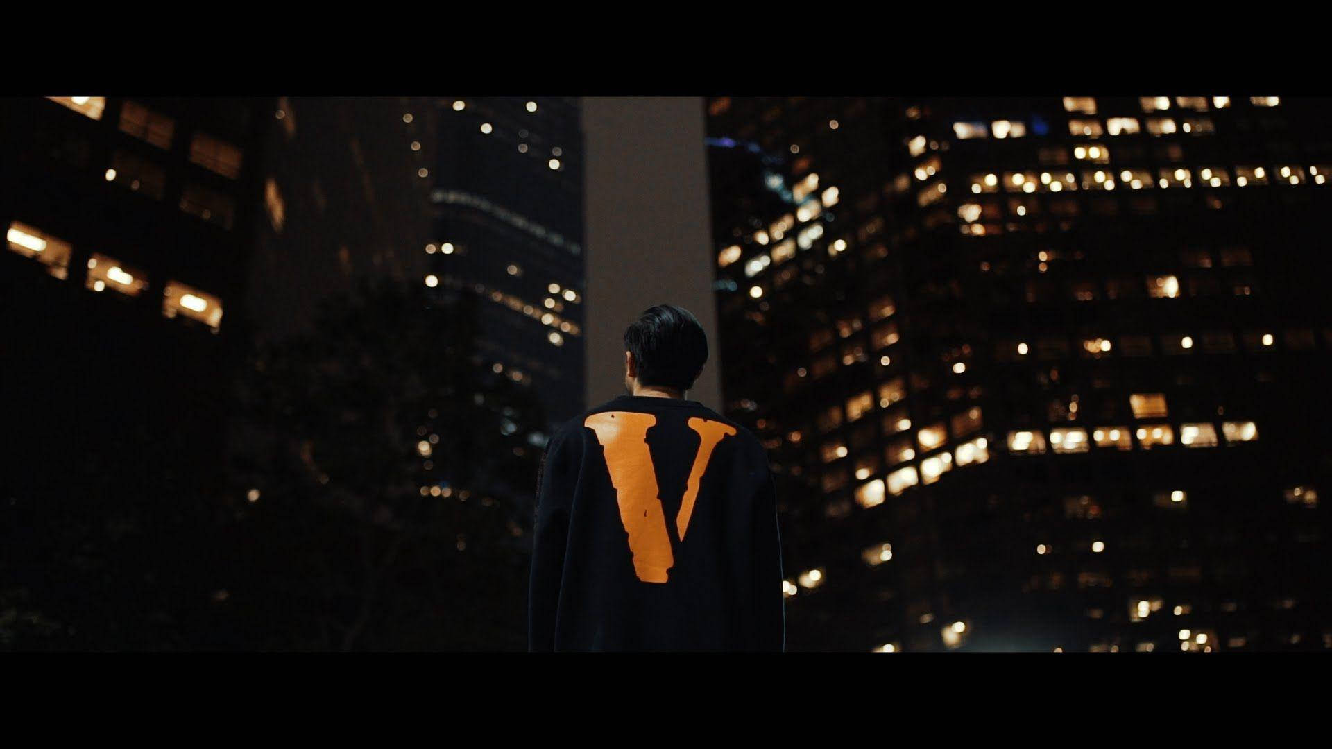 Vlone 桌布 壁紙 — 一個身穿黑色斗篷、帶有發光橙色「V」的人獨自站在夜晚，凝視著燈火通明的摩天大樓城市景觀。