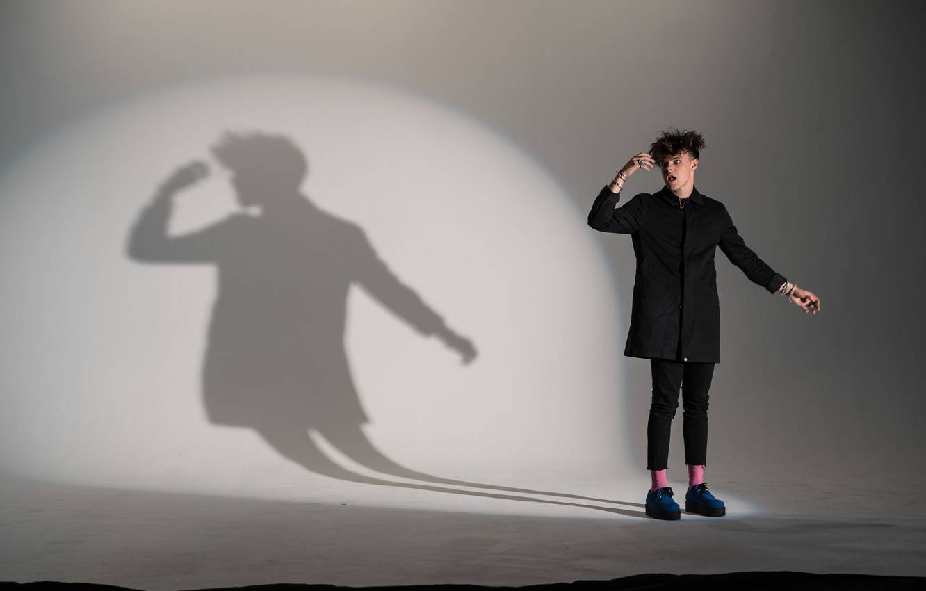 Yungblud 桌布 壁紙 — 一個身穿黑色服裝的人站在白色地板上，身後牆上投射出戲劇性的陰影。