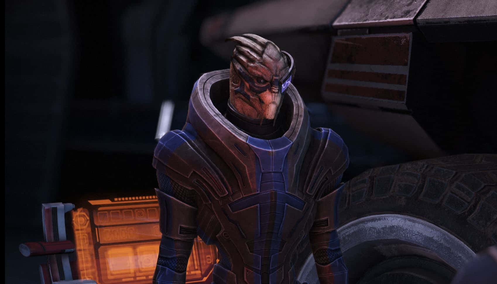 Garrus Vakarian 桌布 壁紙 — 一個身穿黑色盔甲的機械外星戰士，站在光線昏暗的科幻車庫中，附近有發光的橙色面板和一個大輪胎。