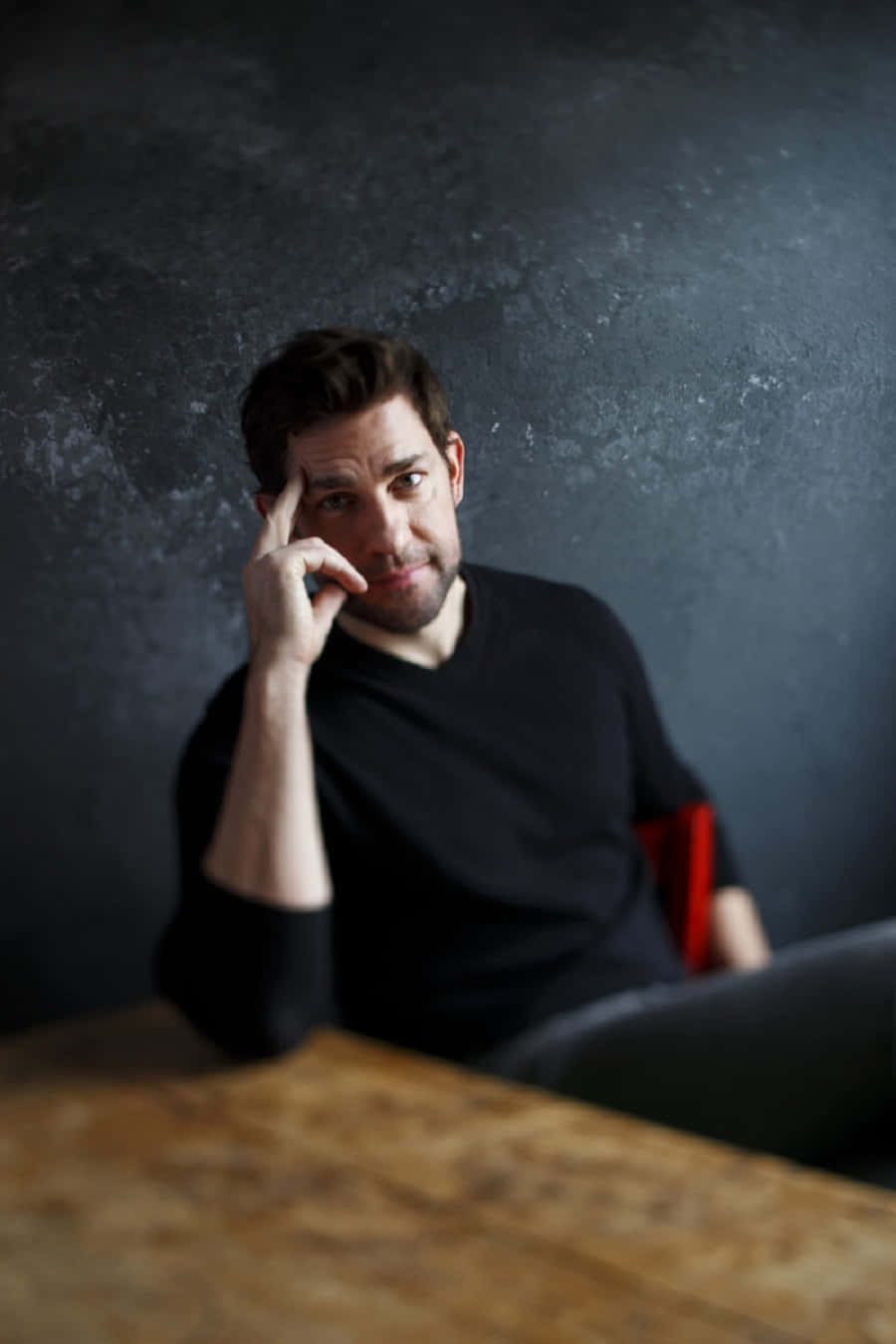 john krasinski 桌布 壁紙 — 一個身穿黑色襯衫的男人坐在木桌前，背景是深色有紋理的牆壁，手放在太陽穴，若有所思。