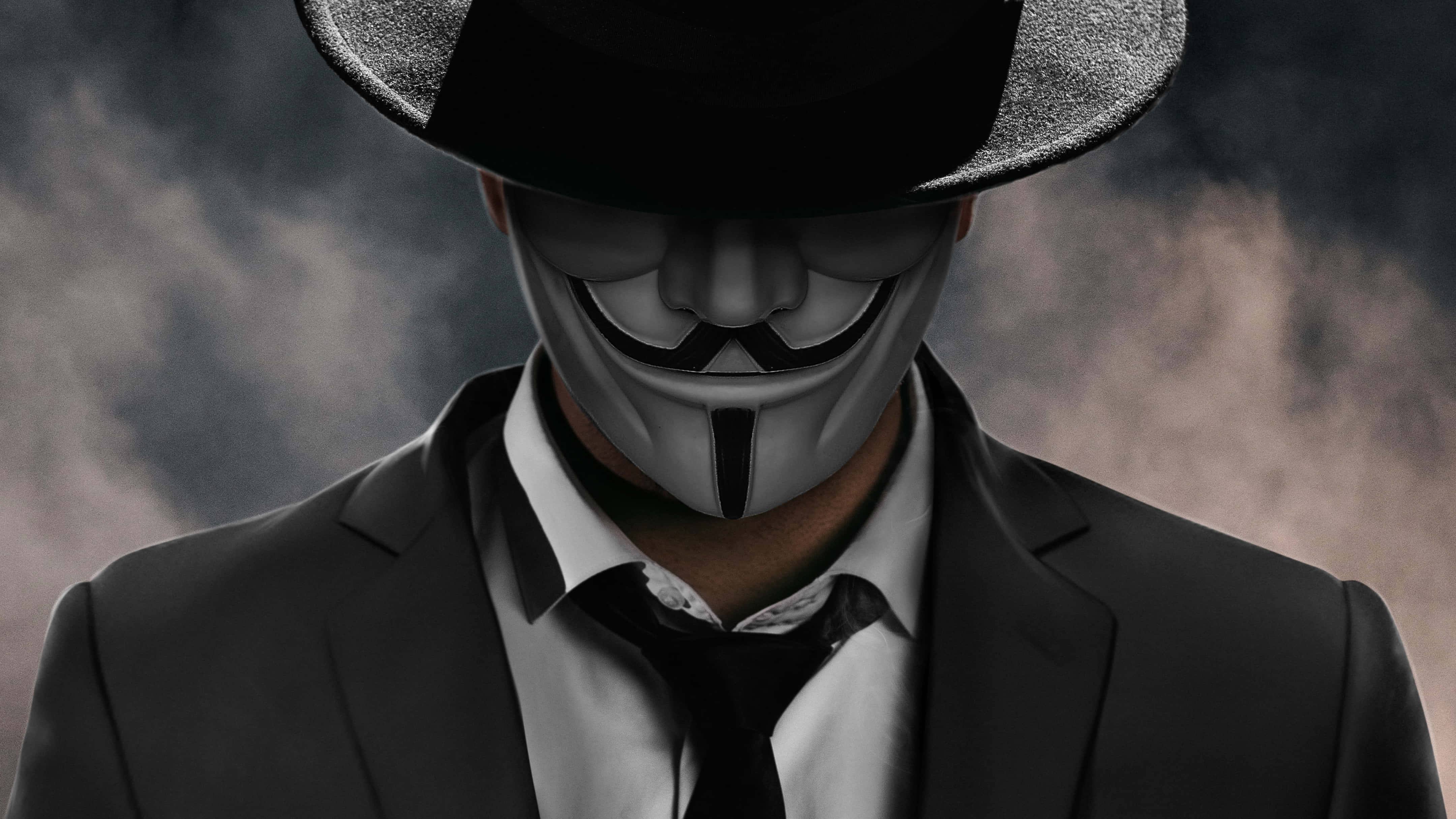 suit 桌布 壁紙 — 一個身穿黑色西裝和軟呢帽的人物，在煙霧彌漫的灰色背景下戴著白色蓋伊·福克斯面具。