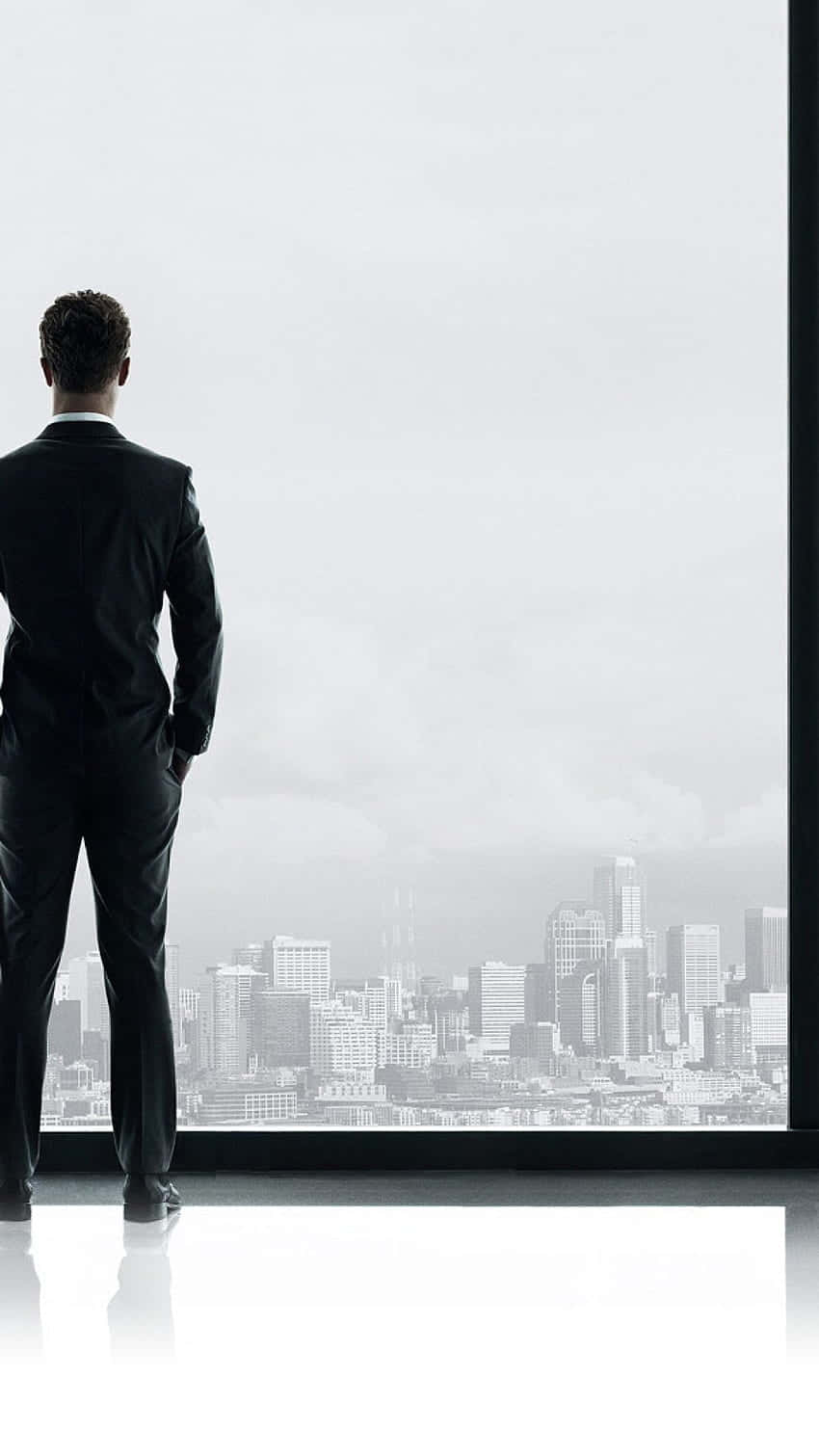 Man In Suit 桌布 壁紙 — 一個身穿黑色西裝的男人獨自站在簡約風格的辦公室裡，透過落地窗凝視著陰天下一片廣闊的城市天際線。