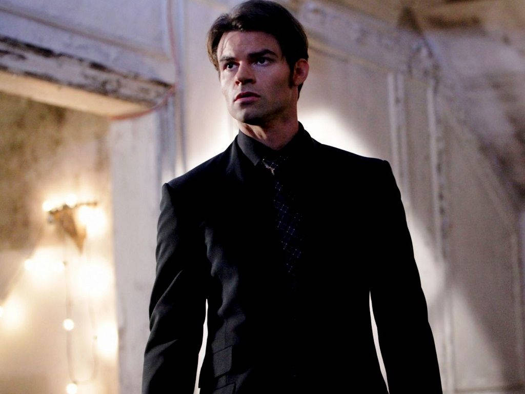 elijah mikaelson 桌布 壁紙 — 一個身穿黑色西裝的男人站在一個光線昏暗、裝飾華麗的走廊裡，身後有溫暖發光的燈光。