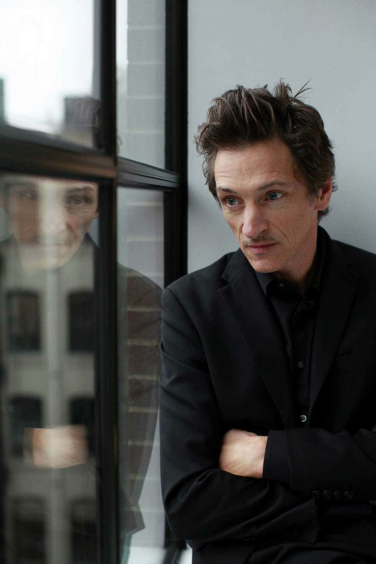 John Hawkes 桌布 壁紙 — 一個身穿黑色西裝的男人雙臂交叉站在窗邊，他的倒影映在玻璃上，若有所思地向外凝望。