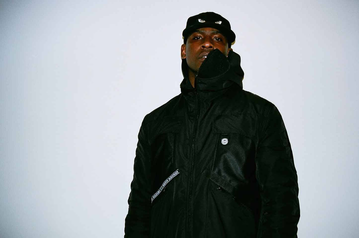 Skepta 桌布 壁紙 — 一個身穿黑色連帽外套和帽子的男人，站在純淺灰色背景前，表情嚴肅。