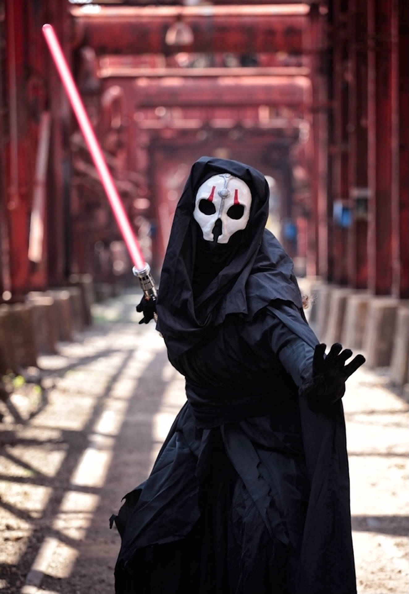 Darth Nihilus 桌布 壁紙 — 一個身穿黑色連帽服裝、戴著白色骷髏面具的人，在陽光照射的木質走廊中揮舞著發光的紅色光劍。