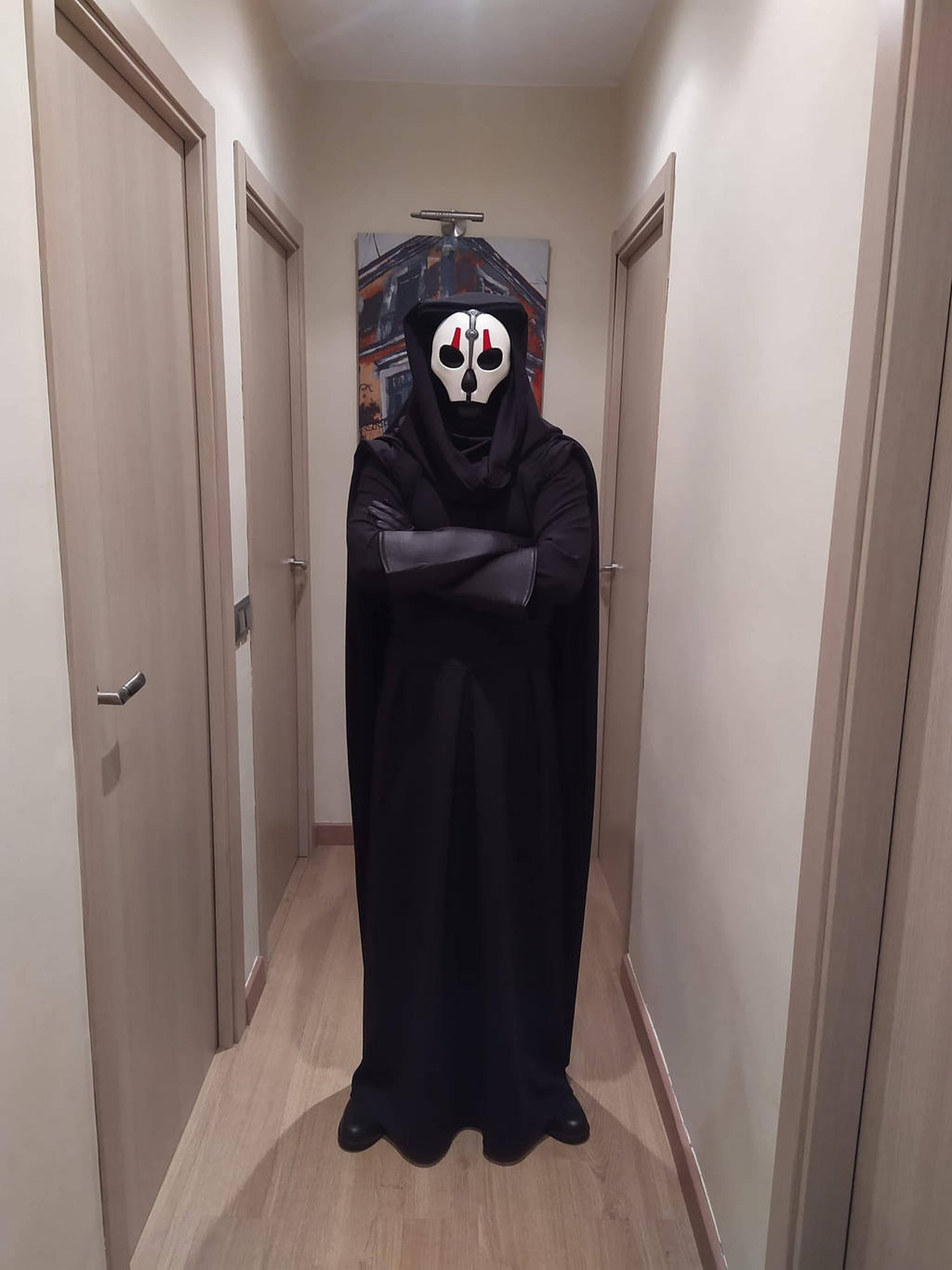 Darth Nihilus 桌布 壁紙 — 一個身穿黑色連帽長袍、戴著紅眼白色面具的人物站在一條有閉合門的狹窄走廊中。
