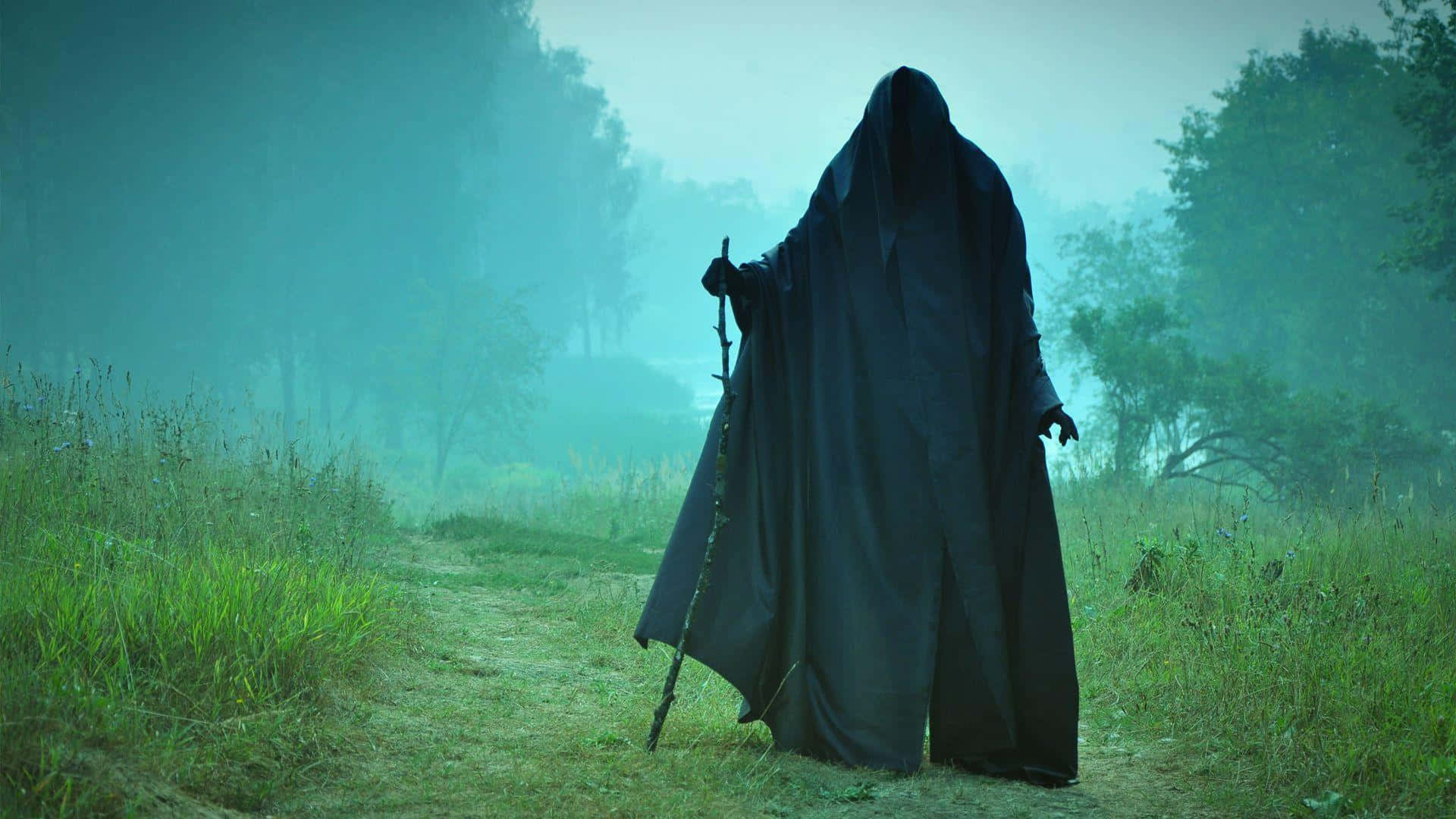 Dark Spirit 桌布 壁紙 — 一個身穿黑色長袍、戴著兜帽的身影，獨自沿著霧氣瀰漫的森林小徑行走，手持一根多節的拐杖。