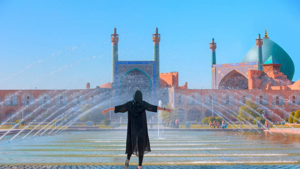 Iran 桌布 壁紙 — 一個身穿黑色長袍的人張開雙臂，站在一座擁有綠松石圓頂和宣禮塔的宏偉清真寺前，在晴朗的藍天下，水柱在他們周圍噴灑。