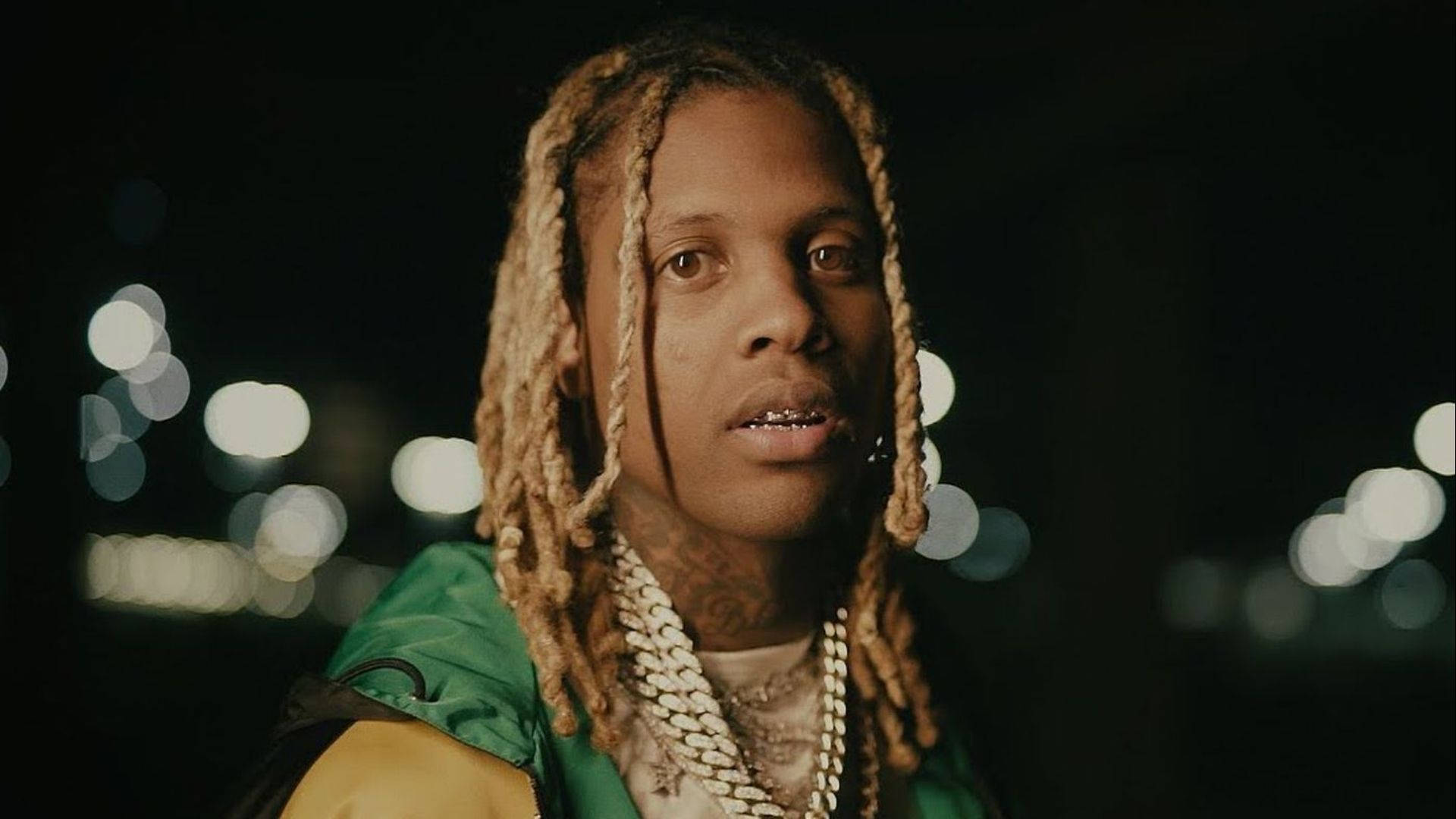 Lil Durk 桌布 壁紙 — 一個辮子頭髮、戴著金鏈的男人，在模糊的夜間城市景觀和發光的散景燈光前專注地凝視。