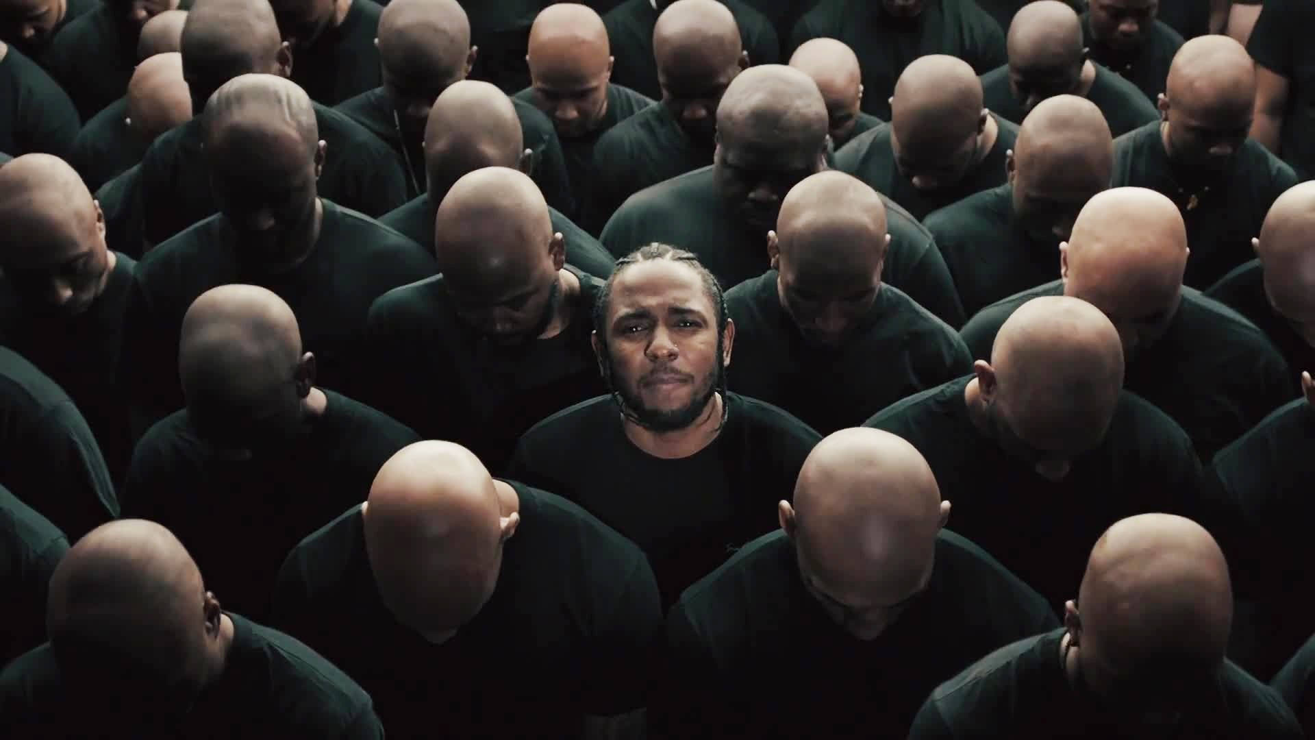 Kendrick 桌布 壁紙 — 一個辮子頭髮的男人獨自向前凝視，被一群穿著黑色襯衫的光頭男人包圍，他們都低著頭。