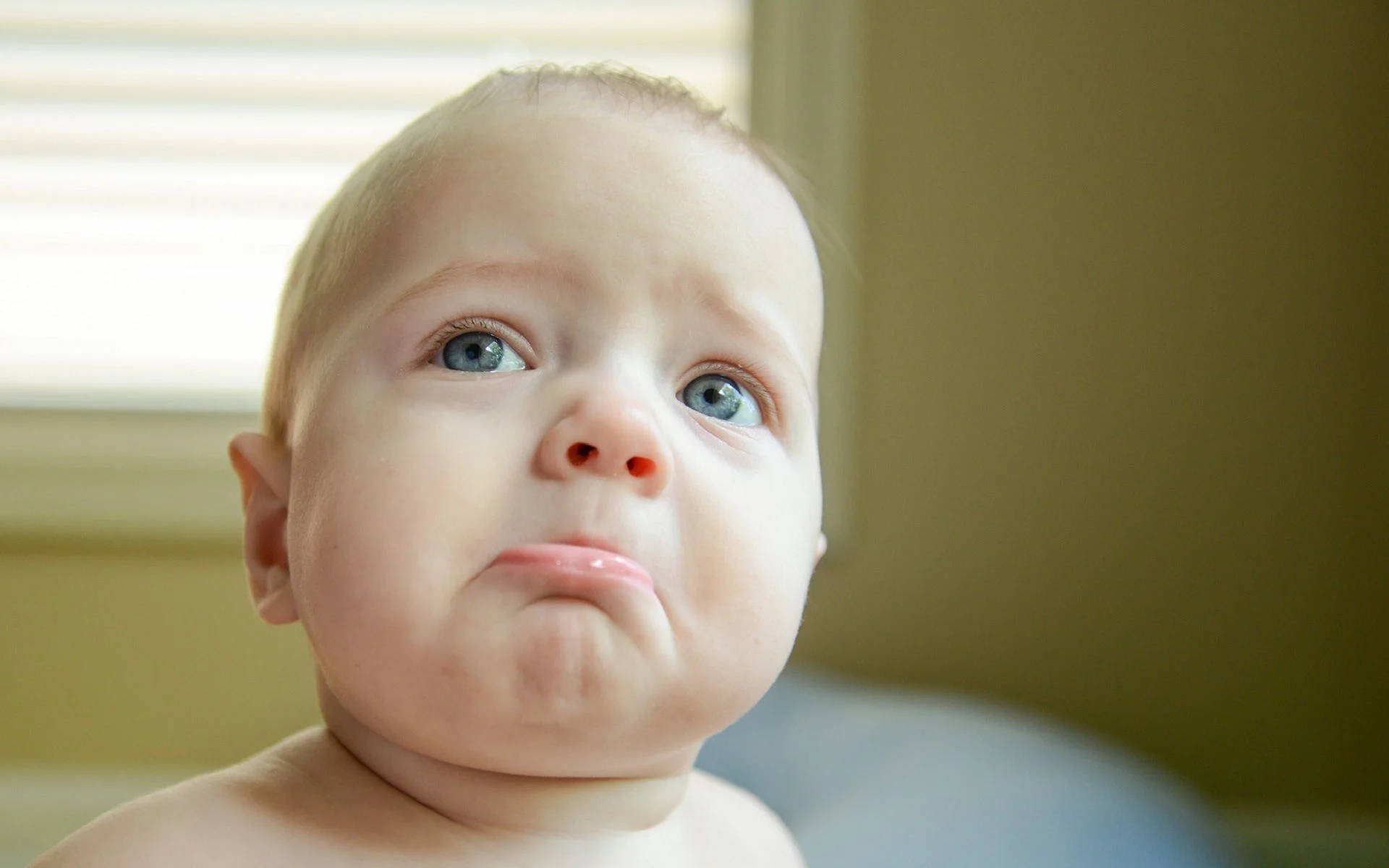 Funny Baby 桌布 壁紙 — 一個近距離拍攝的嬰兒，擁有明亮的藍眼睛和噘嘴的表情，在窗邊向上看。