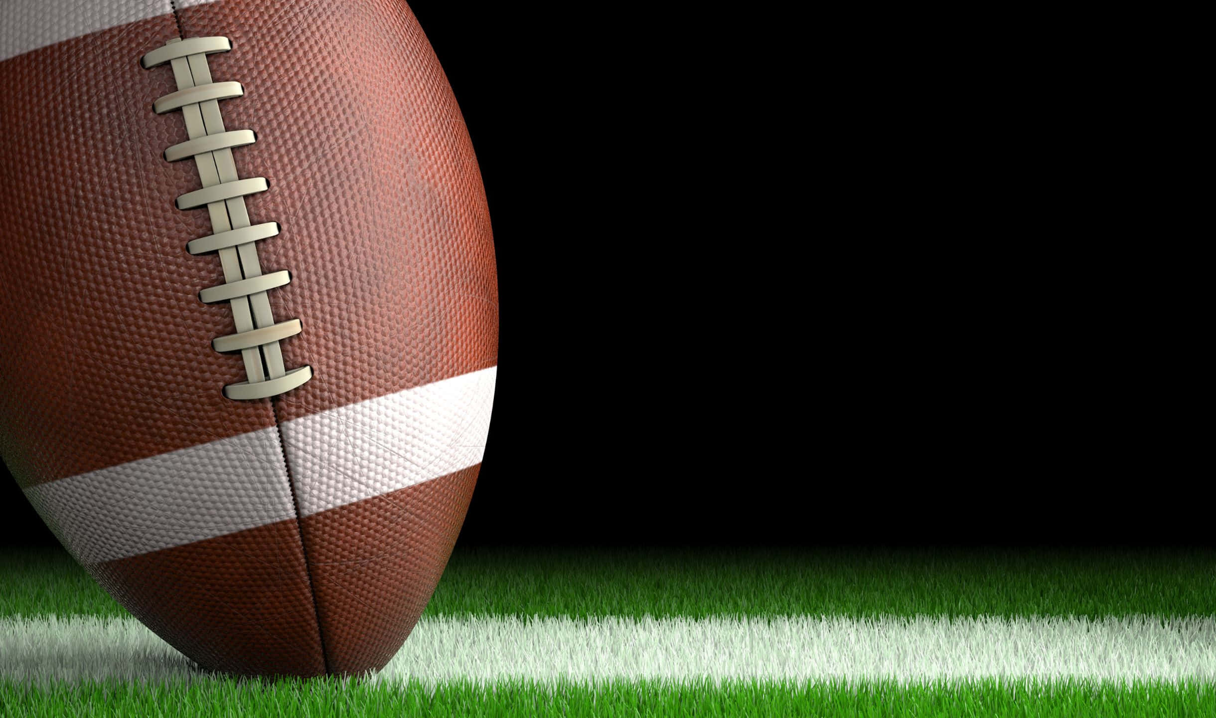 2440x1440 Football 桌布 壁紙 — 一個近距離拍攝的棕色皮革美式足球，帶有白色鞋帶和條紋，放在充滿活力的綠色草地上，背景為黑色。