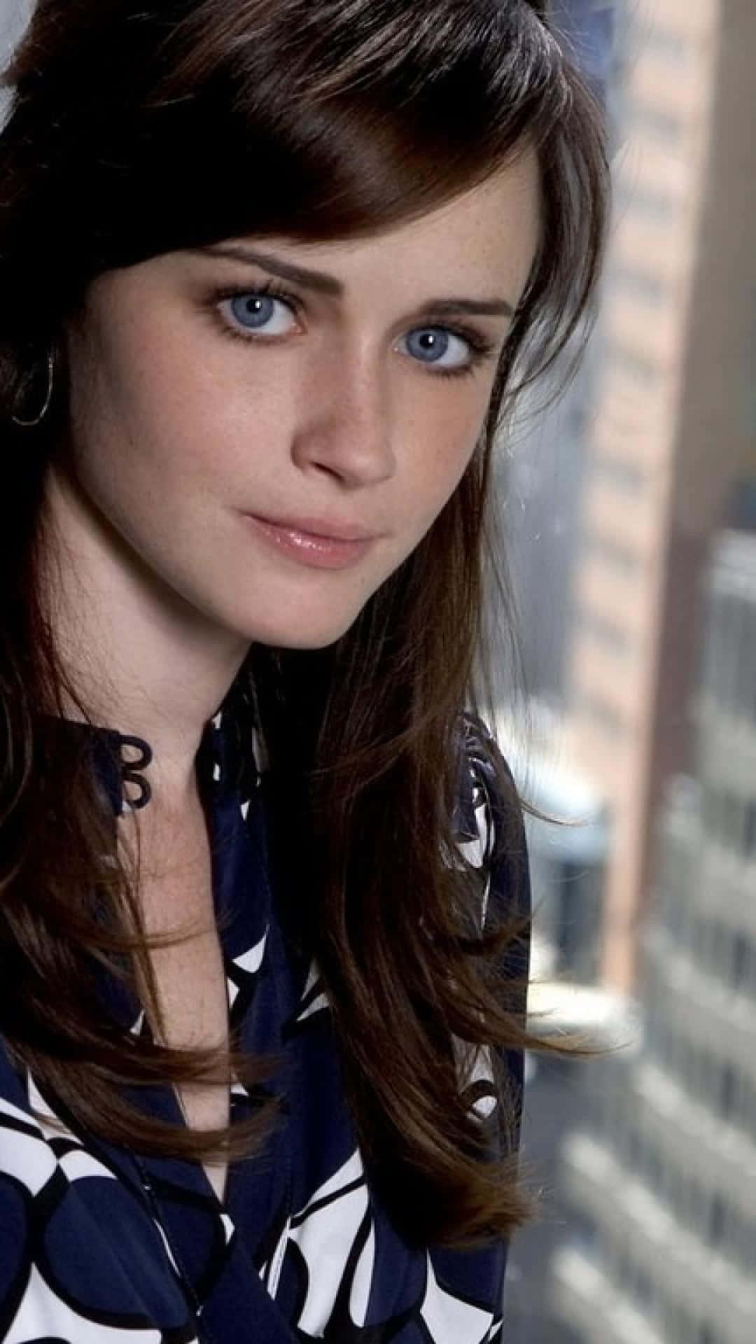 Alexis Bledel 桌布 壁紙 — 一個近距離肖像，一位擁有引人注目的藍色眼睛和棕色頭髮的女性，在模糊的城市景觀背景下直視鏡頭。