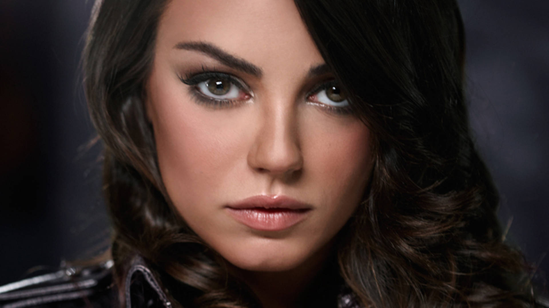 Mila 桌布 壁紙 — 一個近距離肖像，一位擁有深色波浪頭髮和強烈眼神的女性，穿著皮夾克，背景是模糊的深色。