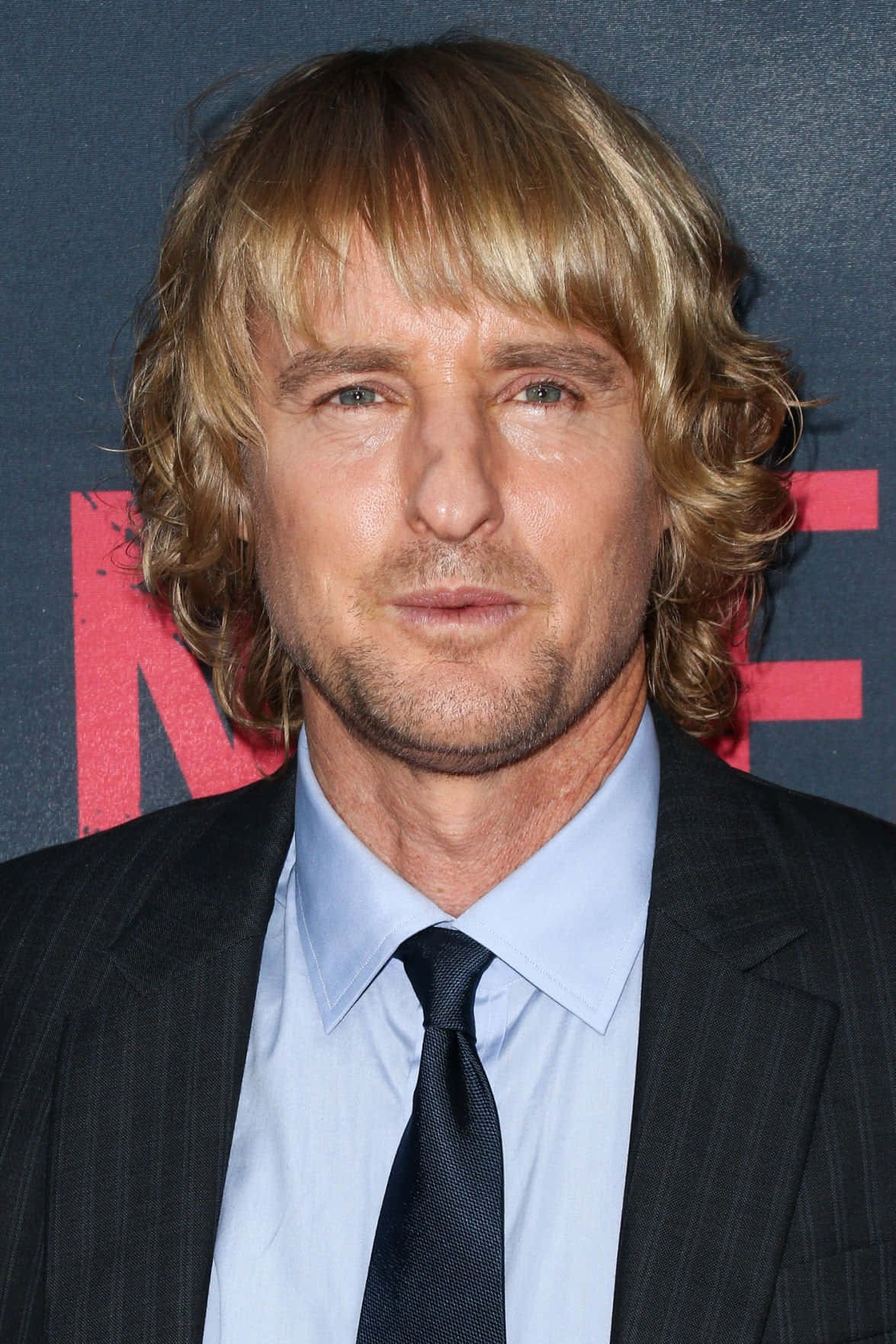 Owen Wilson 桌布 壁紙 — 一個近距離肖像，一個波浪形金髮男子穿著深色西裝和領帶，背景帶有品牌標誌。