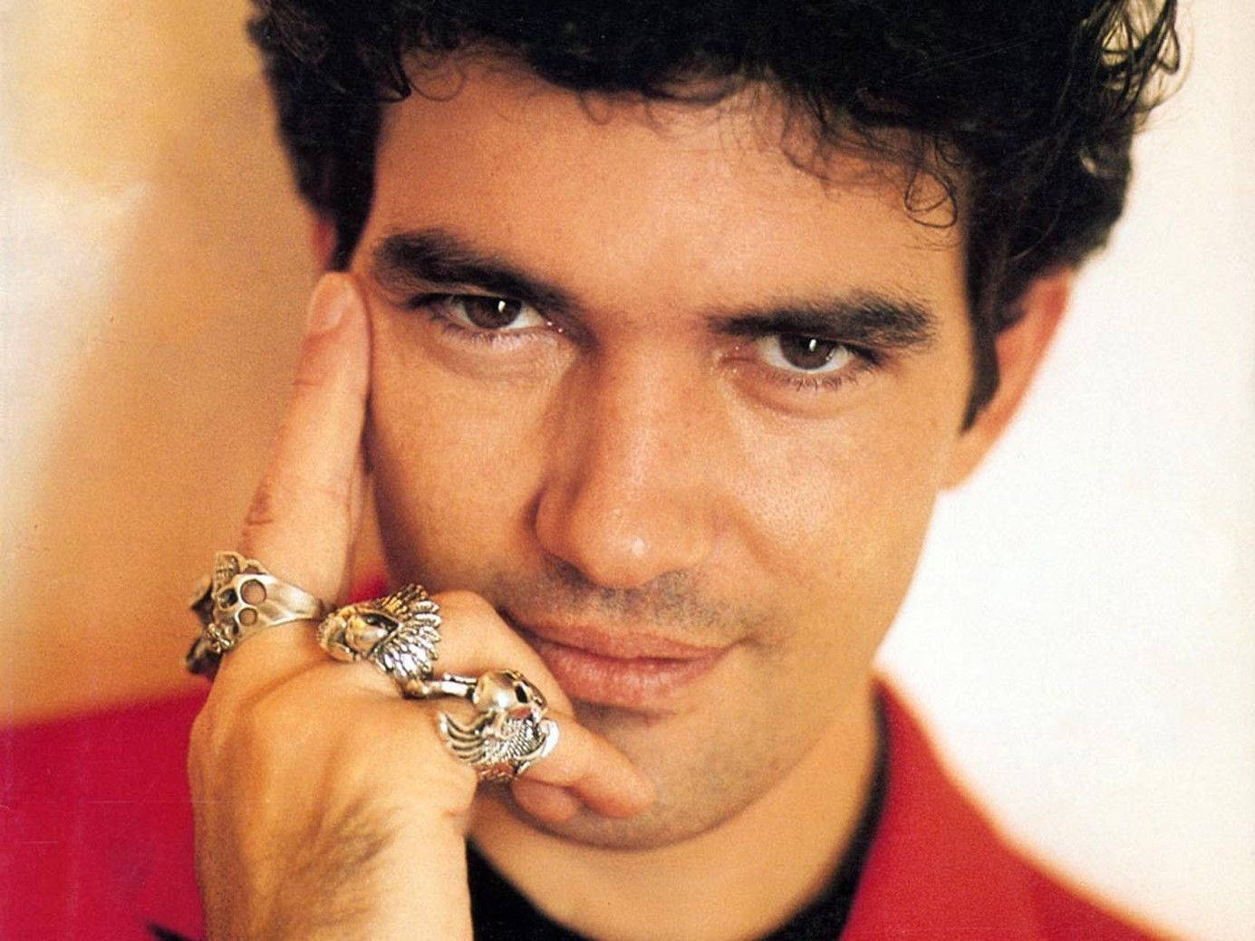 antonio banderas 桌布 壁紙 — 一個近距離肖像，一個深色捲髮、眼神銳利的男人，將下巴擱在一隻戴著多個華麗銀戒指的手上，背景溫暖模糊。