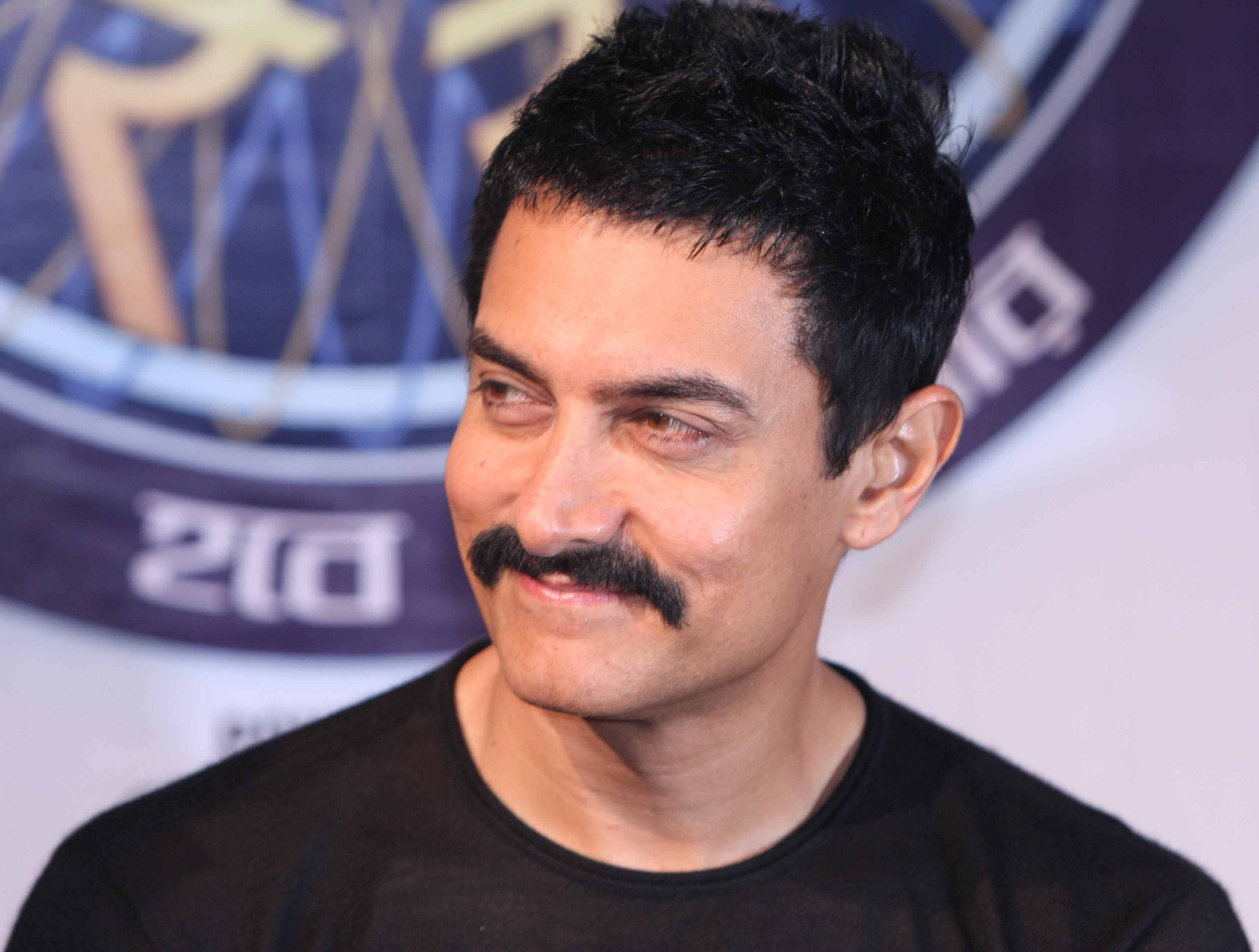 Aamir Khan 桌布 壁紙 — 一個近距離肖像，一個留著濃密鬍鬚的男人穿著黑色T恤，背景模糊帶有品牌標誌。