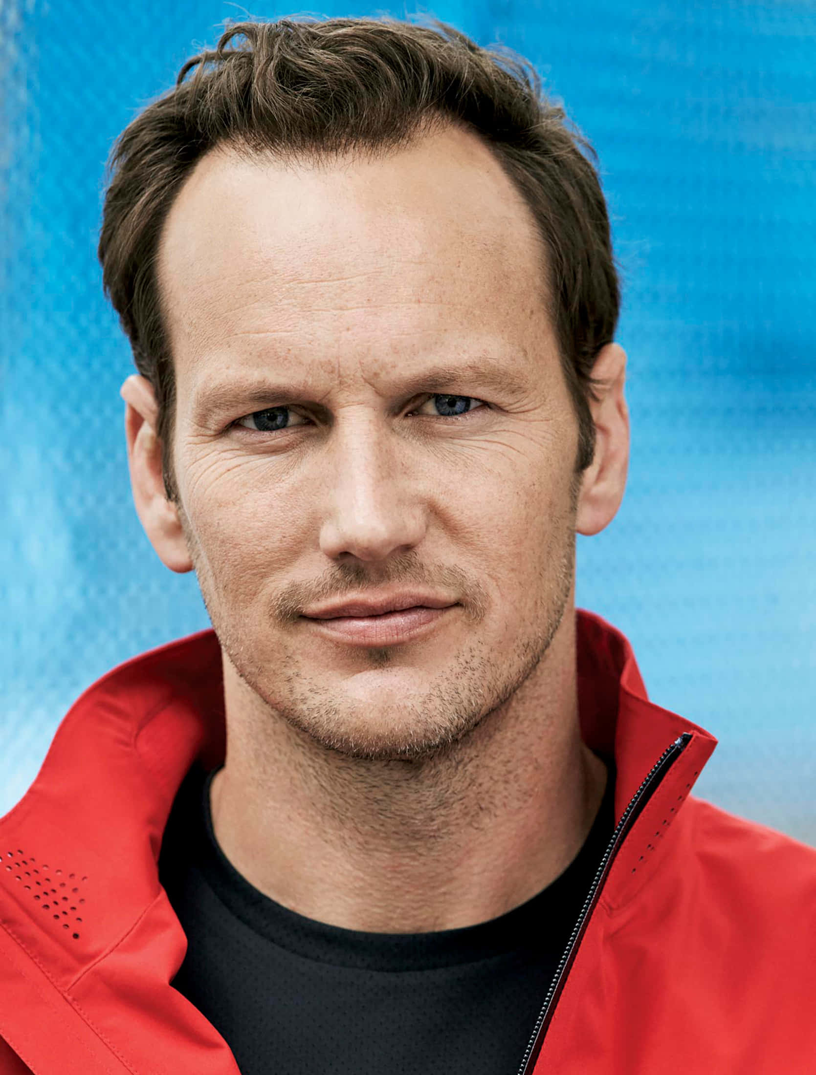 Patrick Wilson 桌布 壁紙 — 一個近距離肖像，一個藍眼睛、鬍渣的男人穿著紅色夾克，背景是帶紋理的藍色。