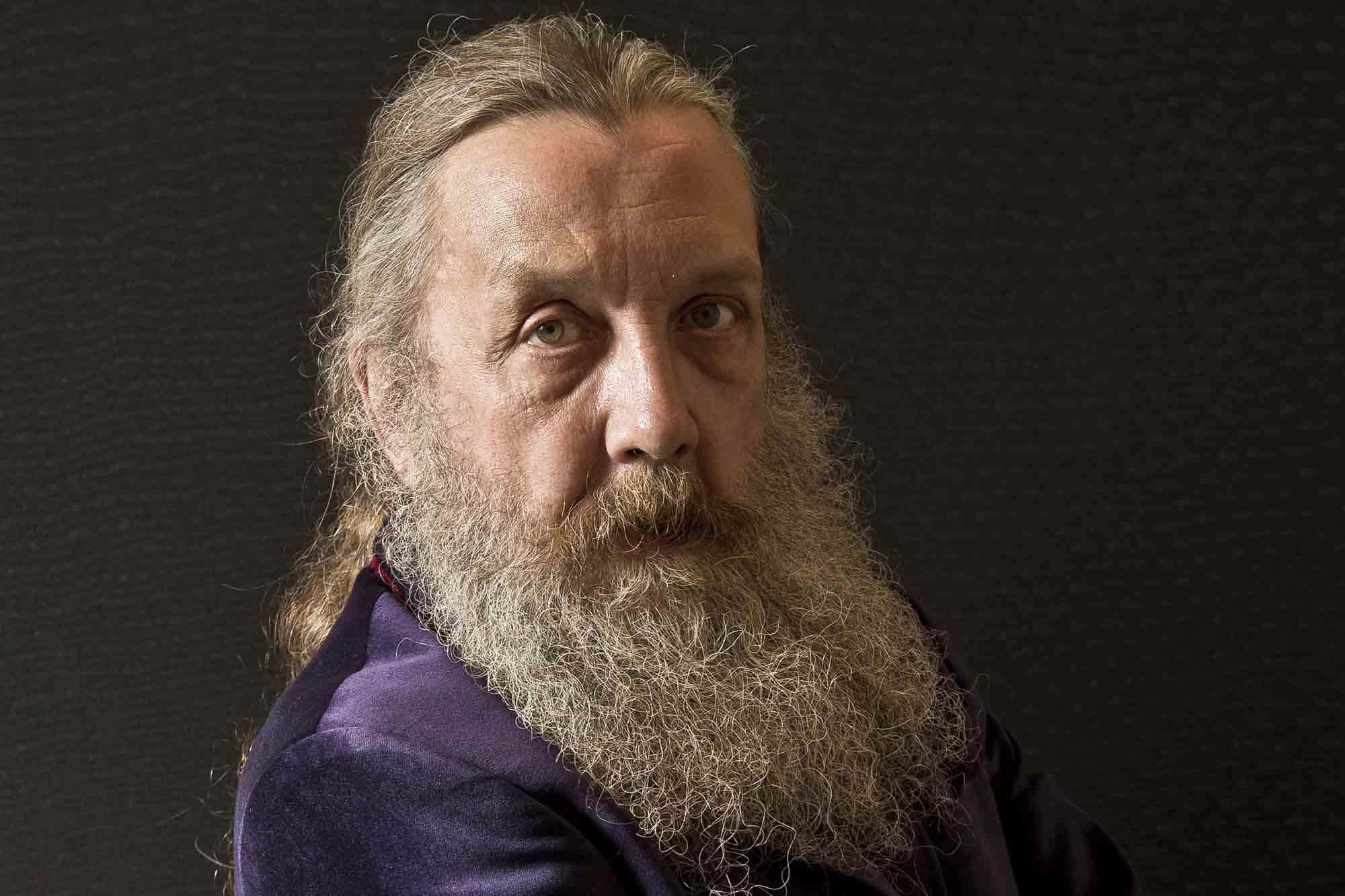 Alan Moore 桌布 壁紙 — 一個近距離肖像，一個長灰髮和滿臉鬍鬚的男人穿著紫色外套，背景是深色有紋理的。