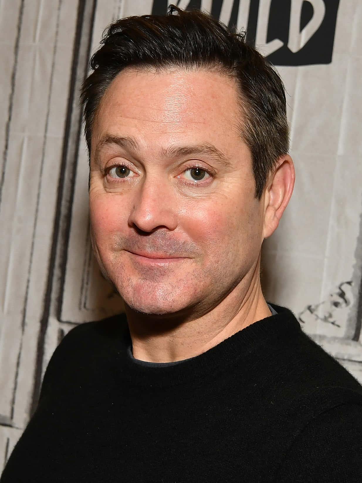 thomas lennon 桌布 壁紙 — 一個近距離肖像，一個黑髮、帶著微妙微笑的男人，穿著黑色毛衣，背景有紋理。