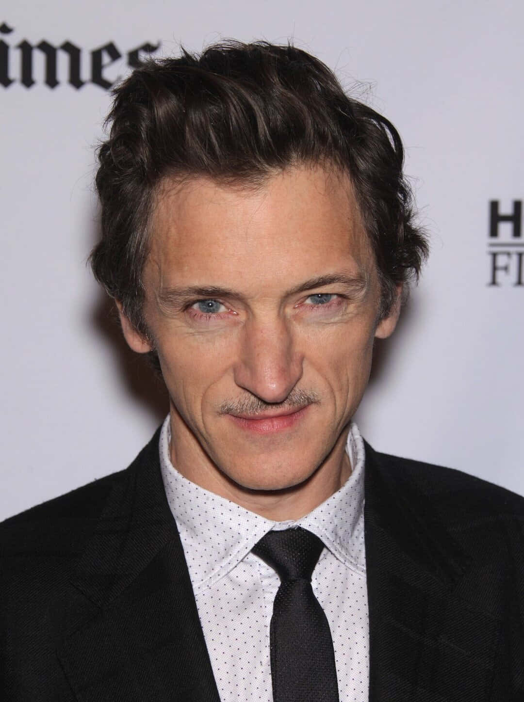 John Hawkes 桌布 壁紙 — 一個近距離肖像，一名藍眼睛、留鬍鬚的男子在正式場合穿著黑色西裝和領帶。