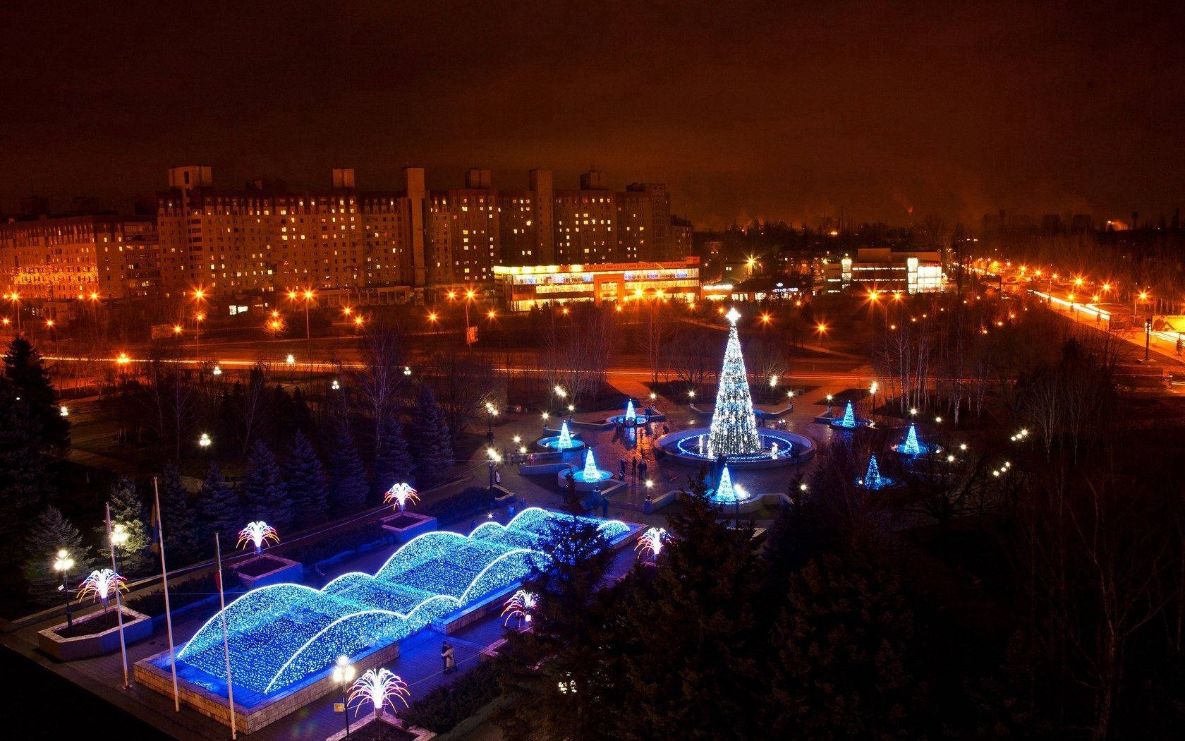 Turquoise 桌布 壁紙 — 一個迷人的夜間城市公園，由波浪形狀的藍色燈光和一棵高聳的聖誕樹照亮，周圍是光禿禿的樹木和遠處的公寓大樓。